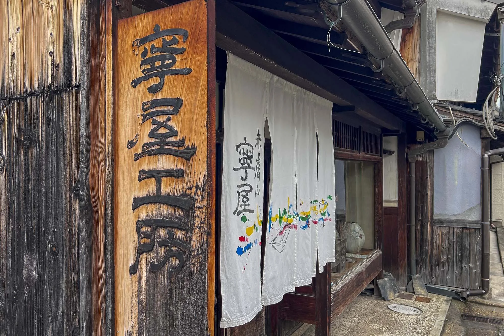 Traditional Japanese storefront with wooden exterior, white noren curtain, colorful painted characters, and a wooden sign with Japanese kanji.