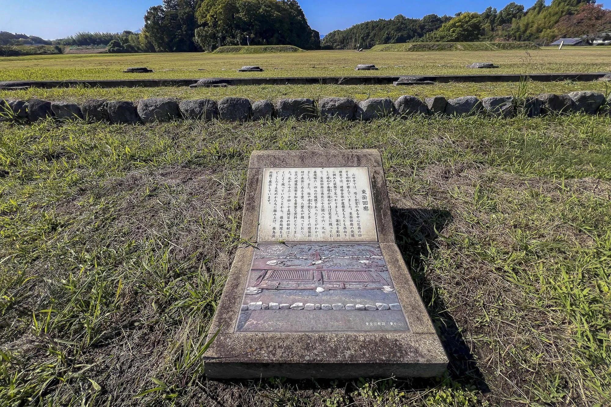 A historical plaque with Japanese text and an illustration, situated in a grassy field with a circular stone arrangement and trees in the background.