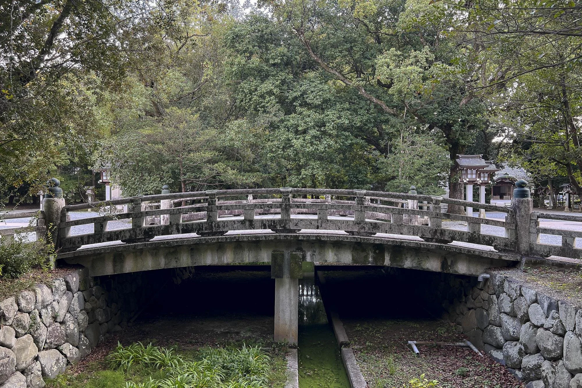 A stone bridge over a narrow water canal in a green, wooded area with trees and traditional Japanese structures in the background.