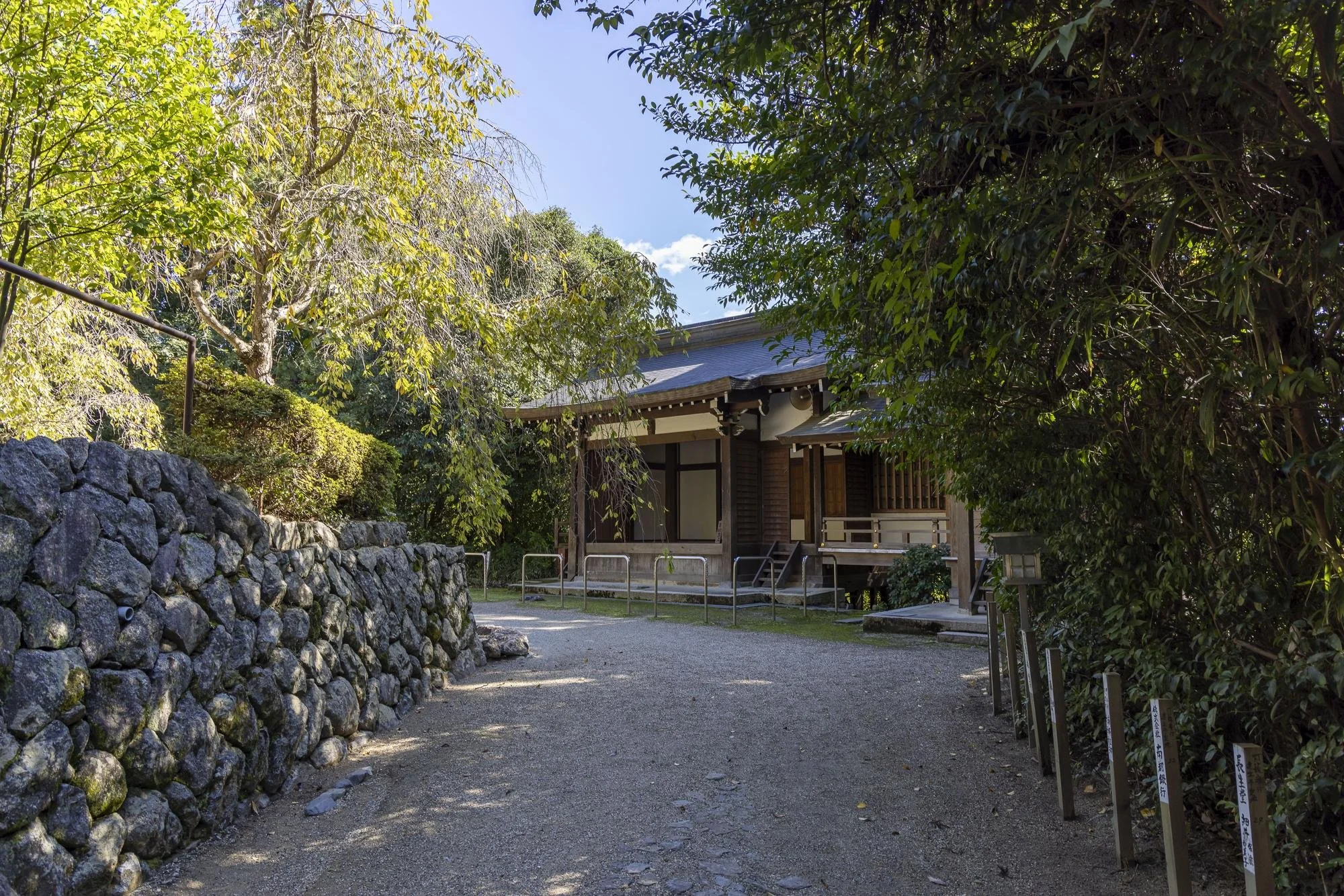 A traditional Japanese wooden building with a sloped roof surrounded by trees and greenery, with a gravel path leading up to it.