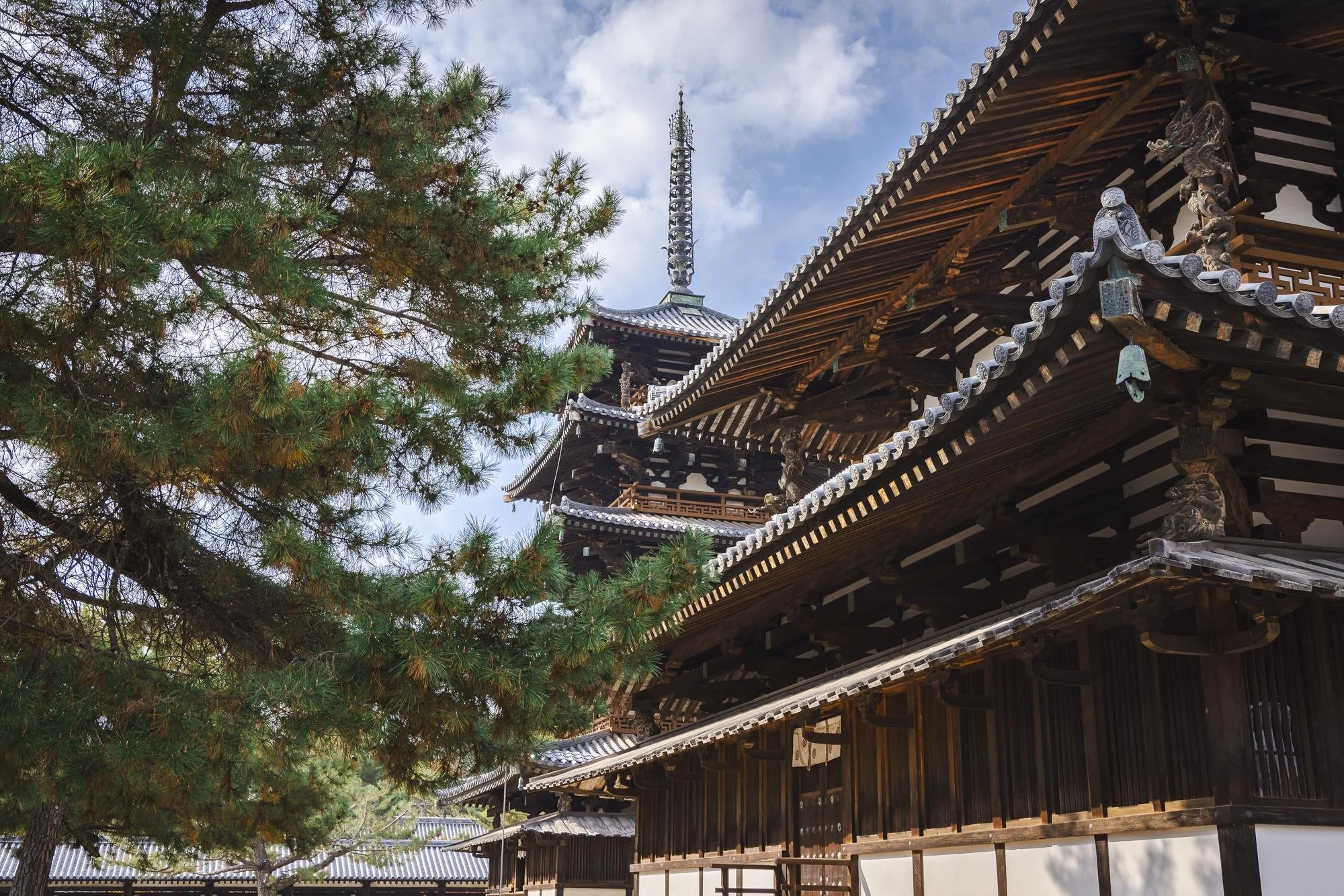 Traditional Japanese temple with wooden architecture, a tiled roof, and a pagoda spire in the background, surrounded by green pine trees and a blue sky with clouds.