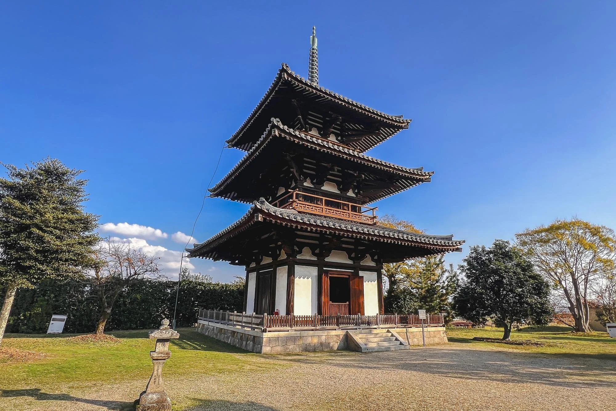 Traditional Japanese pagoda with multiple tiers, dark wooden structure, white walls, set in a park with trees and clear blue sky.