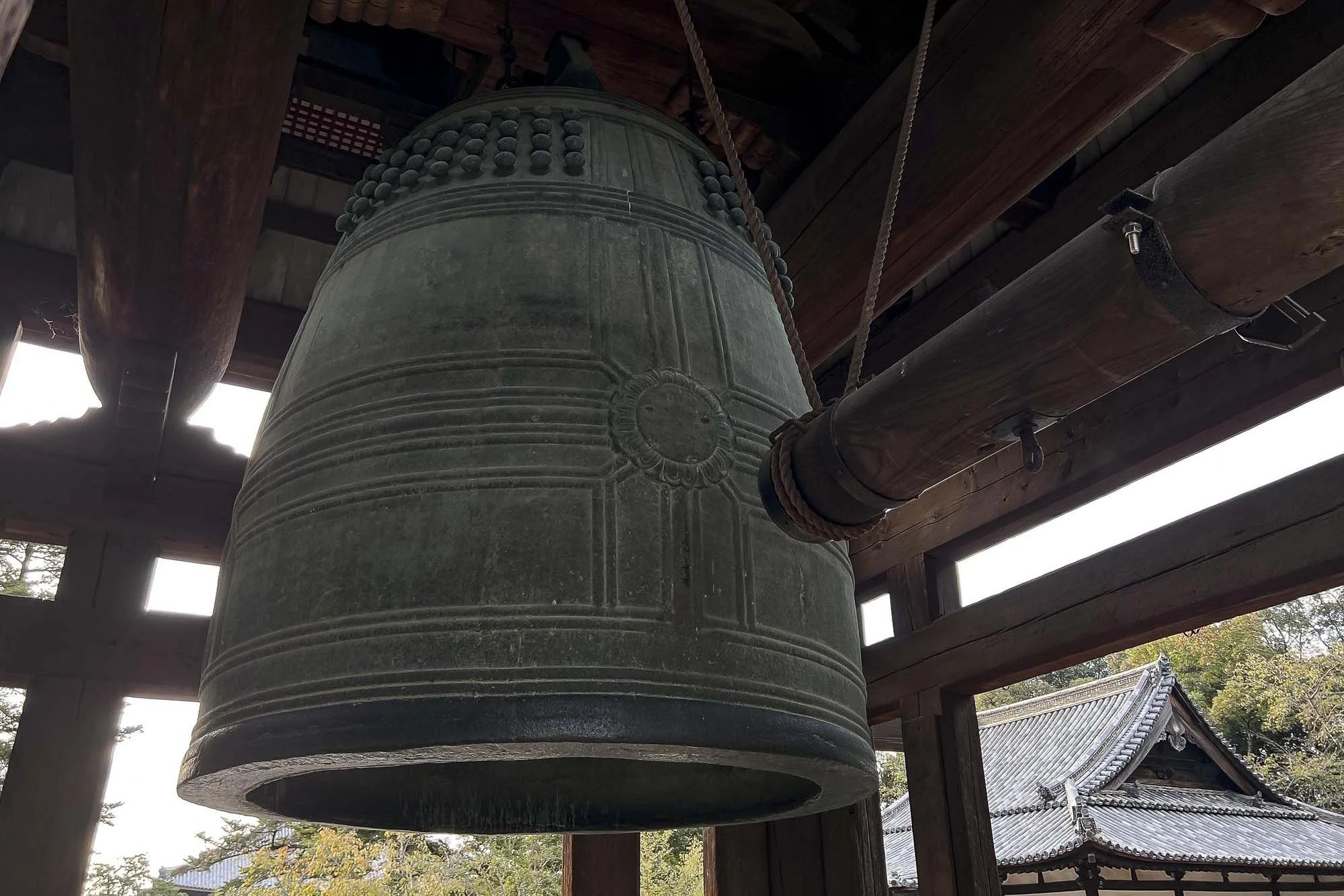 Large traditional bronze bell hanging inside a wooden structure at a Japanese temple with a distant roof visible outside.