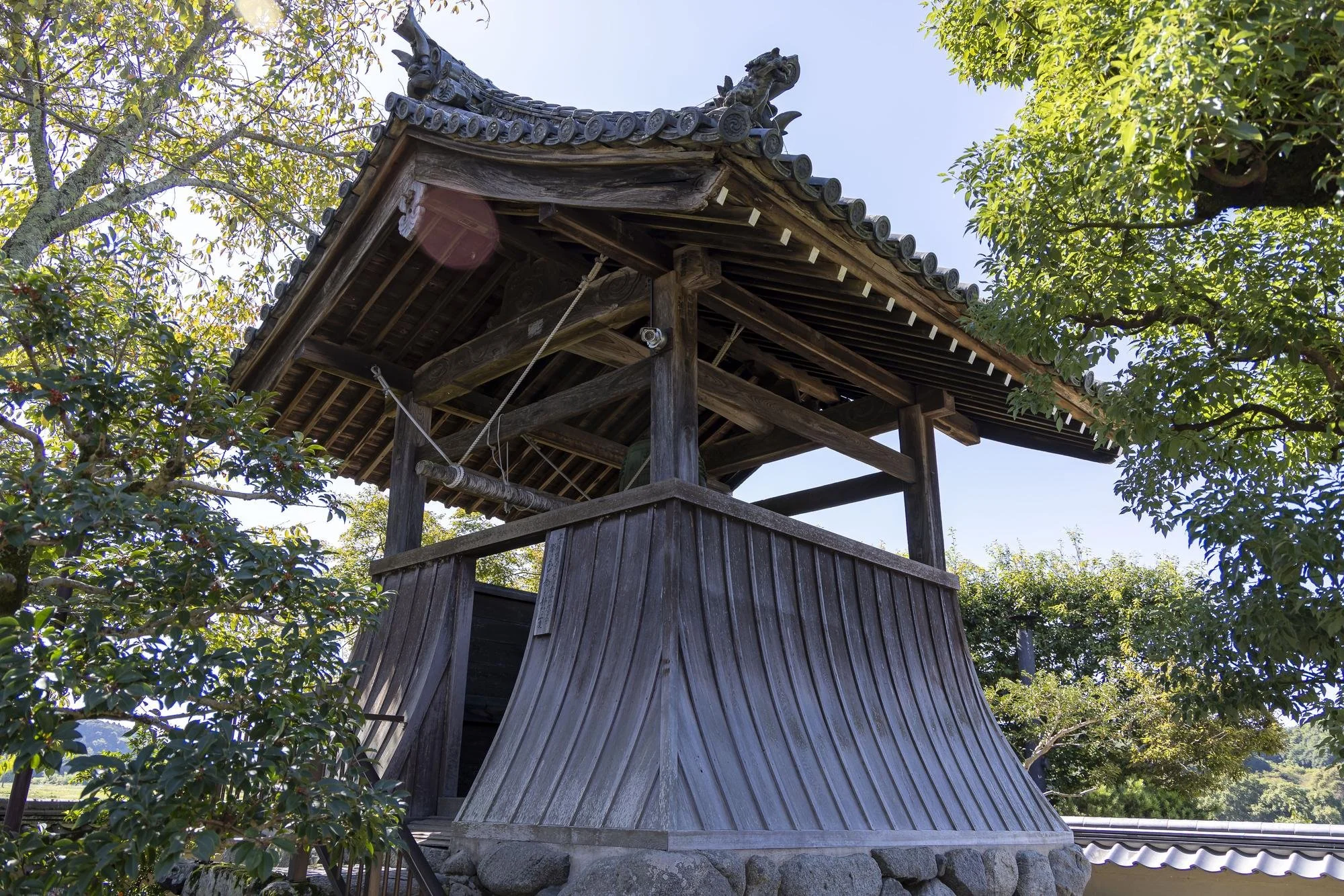 Wooden bell tower with traditional Japanese architectural style, surrounded by trees and clear sky.