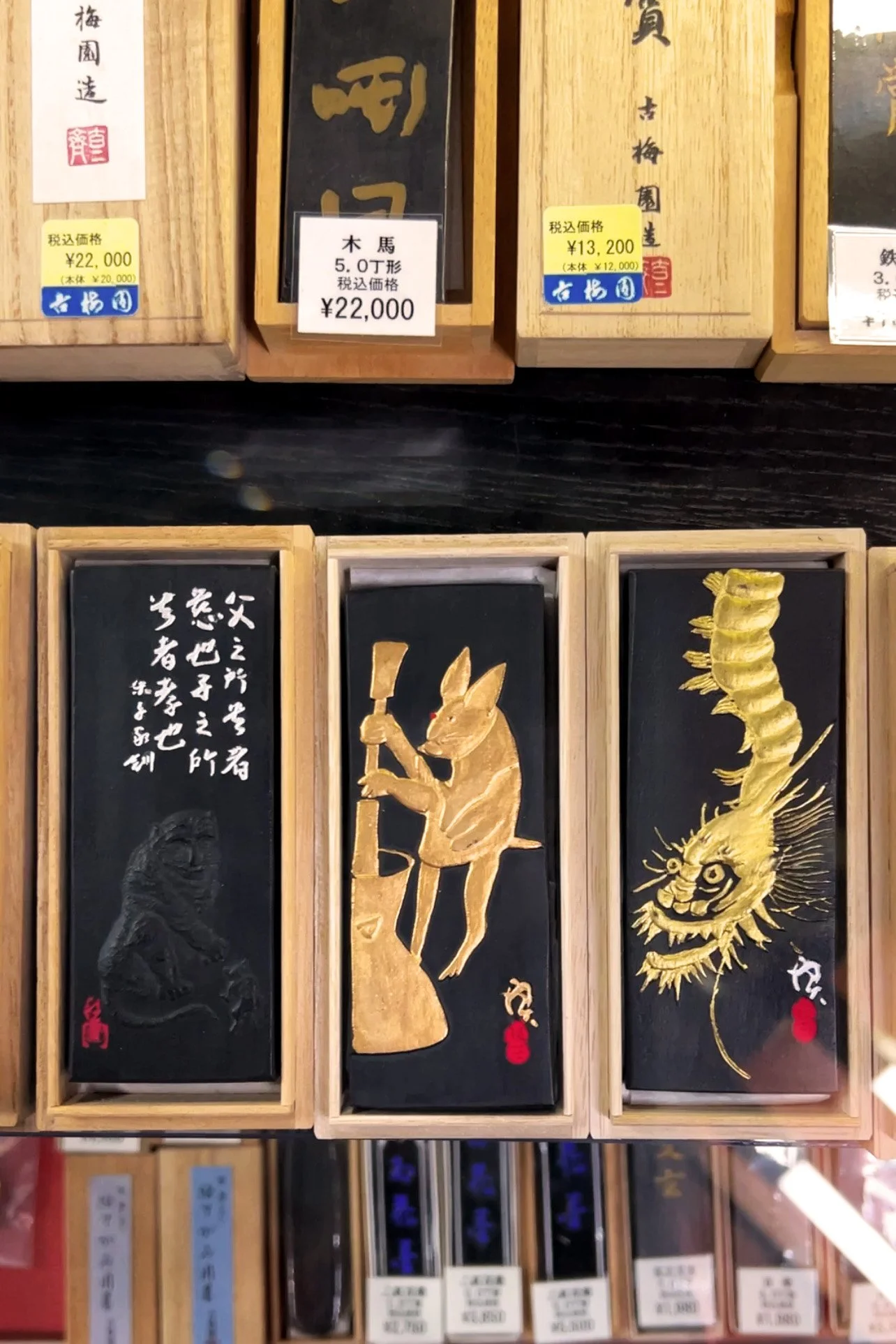 Three wooden boxes with black backgrounds, gold painted images of a fox holding an axe, a dragon, and some Japanese characters layered with paper labels, set amongst other boxed items.