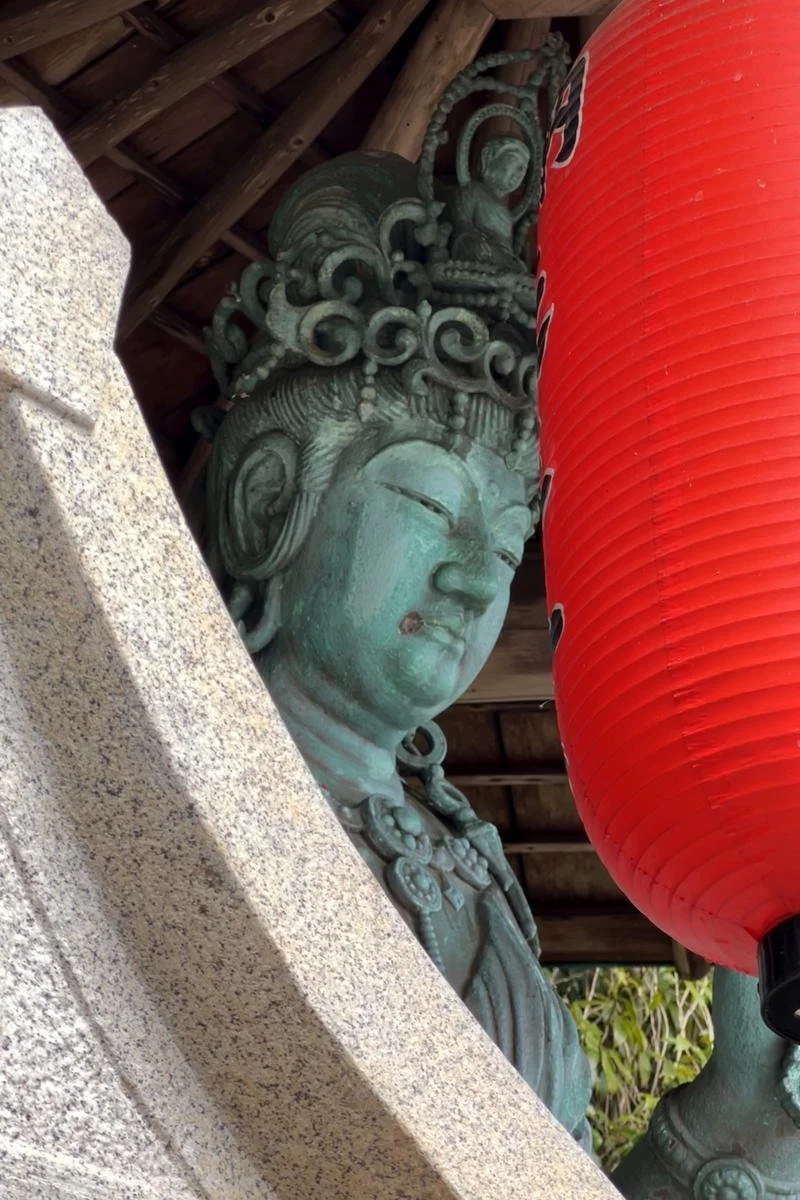 A sculpture of a deity or spiritual figure partially obscured by a large red lantern, located under a wooden roof.