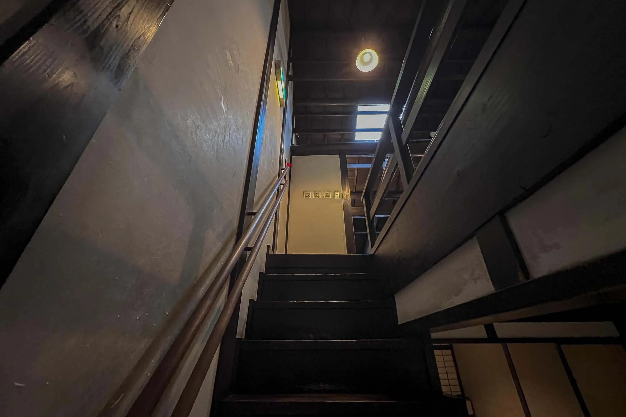 Interior view of a staircase leading up to a second floor with dark wood railing, wooden stairs, and a window allowing natural light, with ceiling light and electricity switches on a wall.