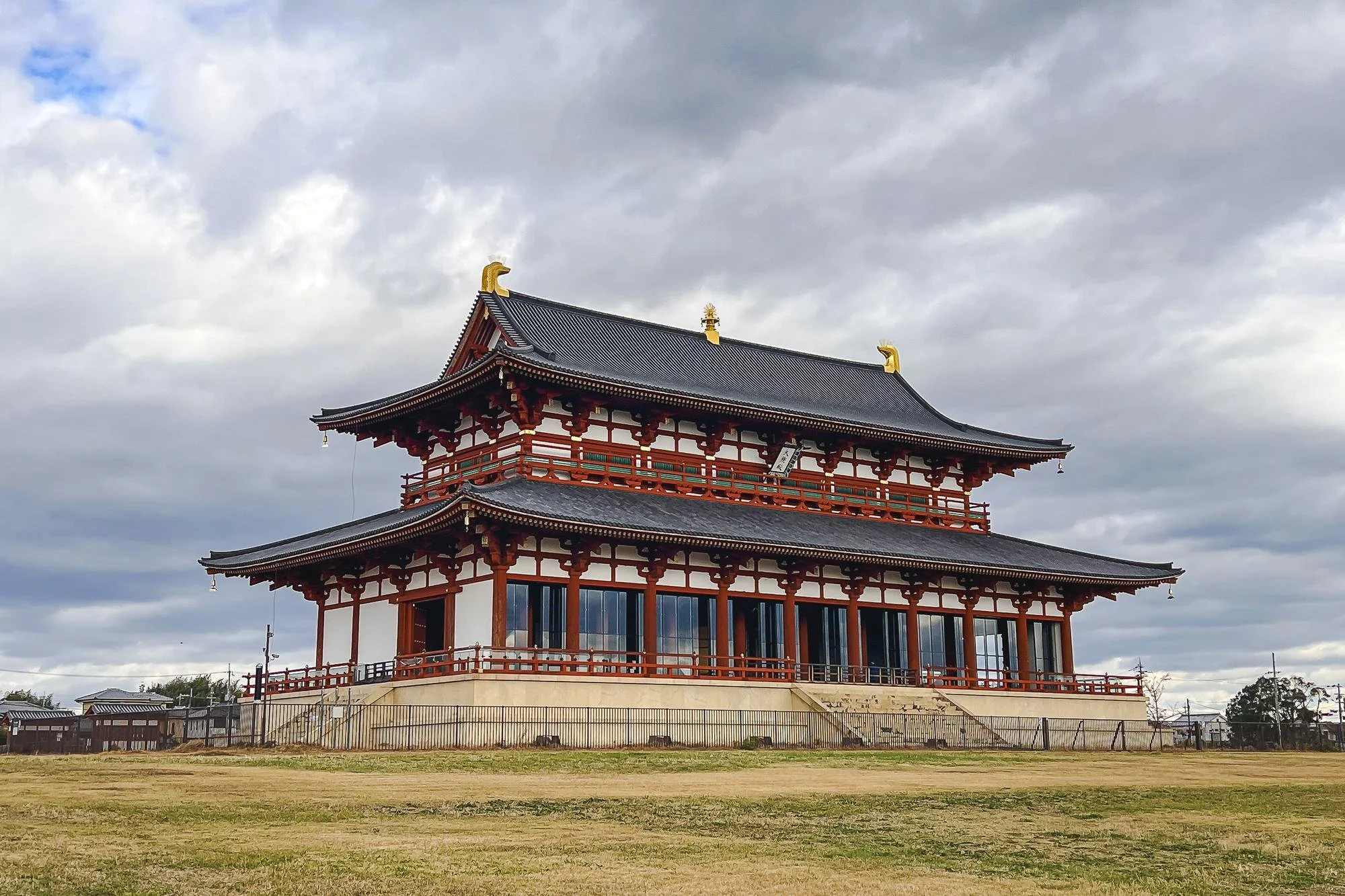 Traditional Japanese temple with red wooden structure, gray tiled roof, and decorative gold accents, set against a cloudy sky and open grassy area.