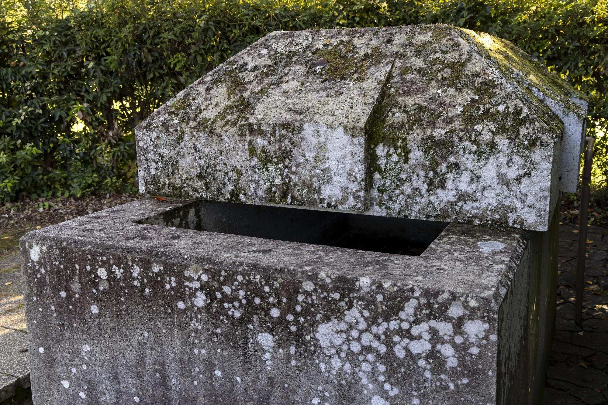 An old stone water fountain with a rectangular basin and a sloped roof, covered in moss and lichen, situated outdoors with green shrubbery in the background.