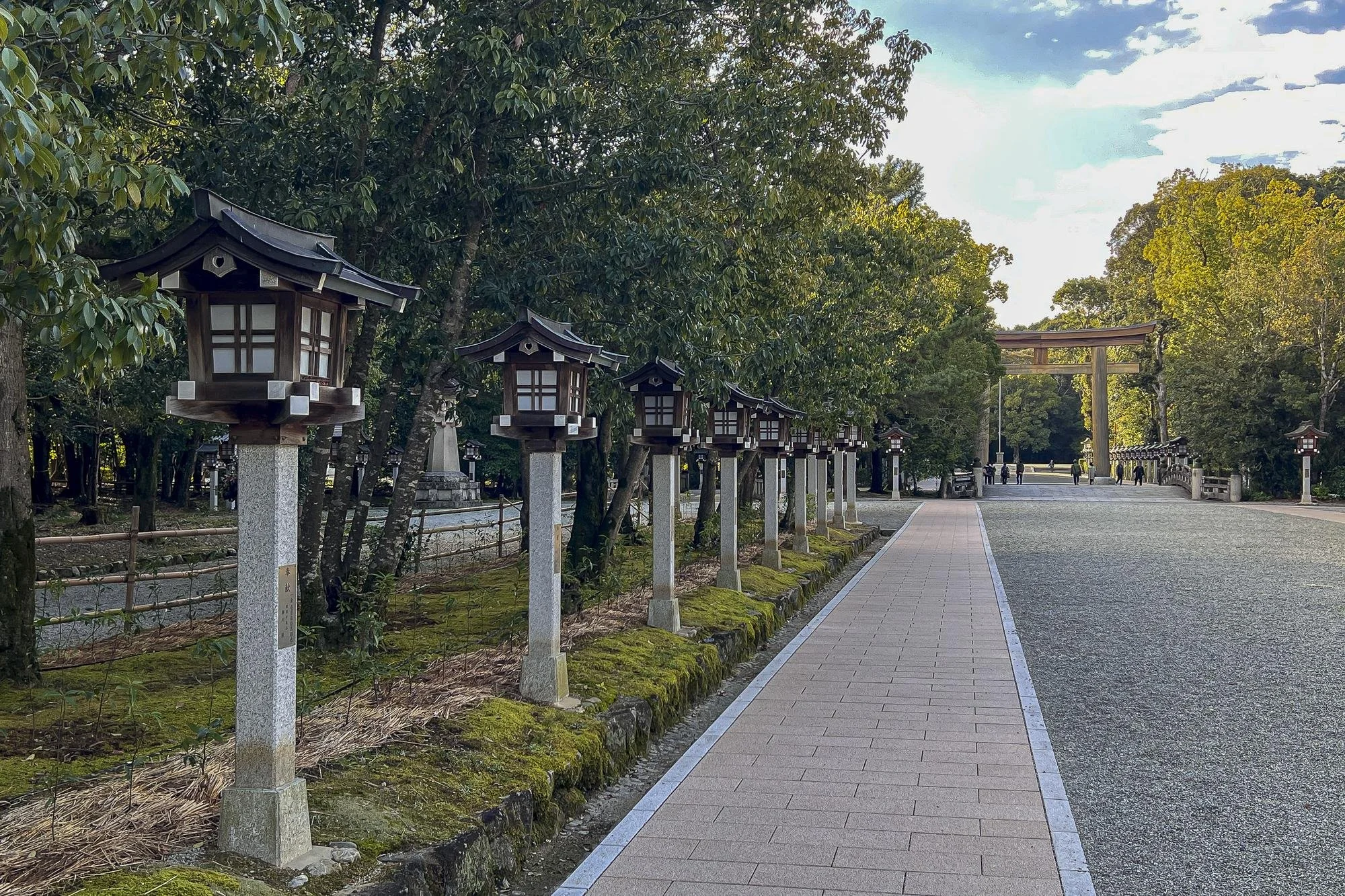 Pathway leading to a traditional Japanese torii gate, flanked by small lanterns on stone pillars, amidst lush green trees.