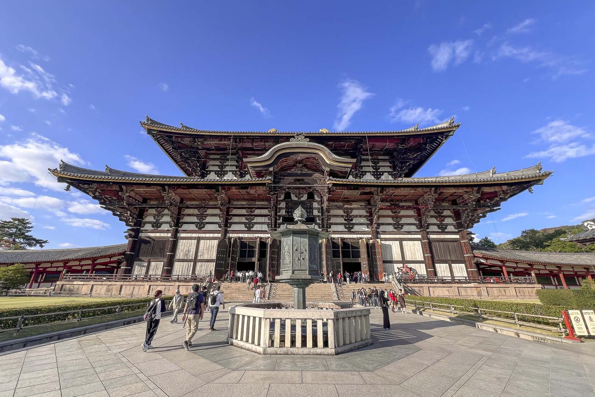 A traditional Japanese temple with tiered, curved rooflines surrounded by visitors, under a blue sky with some clouds.