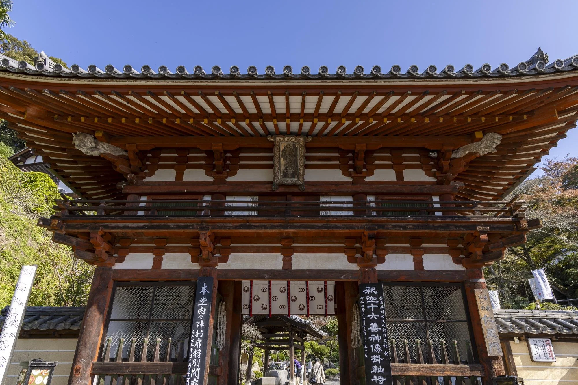 Traditional Japanese temple gate with wooden structure, with a blue sky above and trees on the sides.