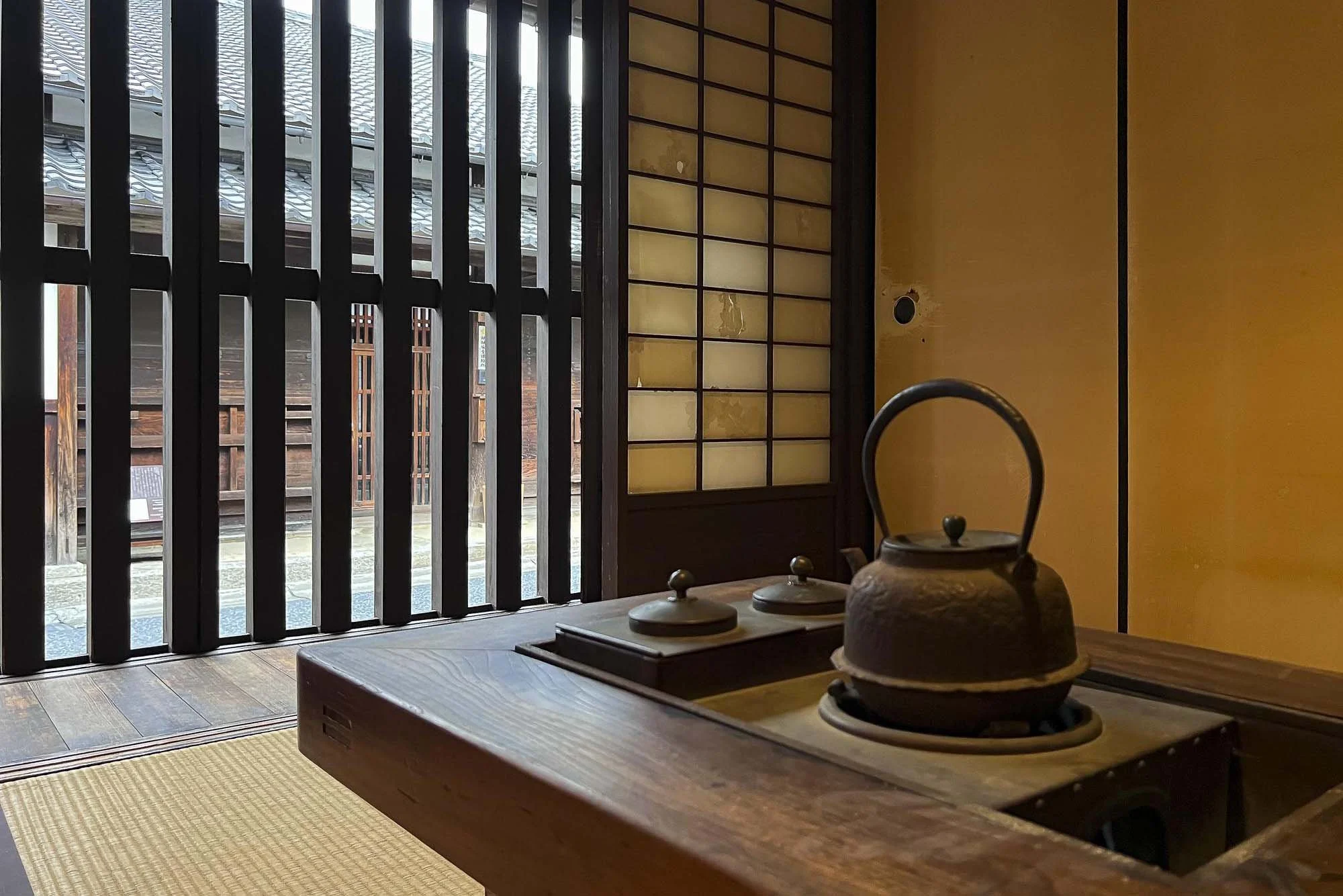 Traditional Japanese room with a kettle on a stove, shoji sliding door, tatami mat flooring, and wooden slat window.