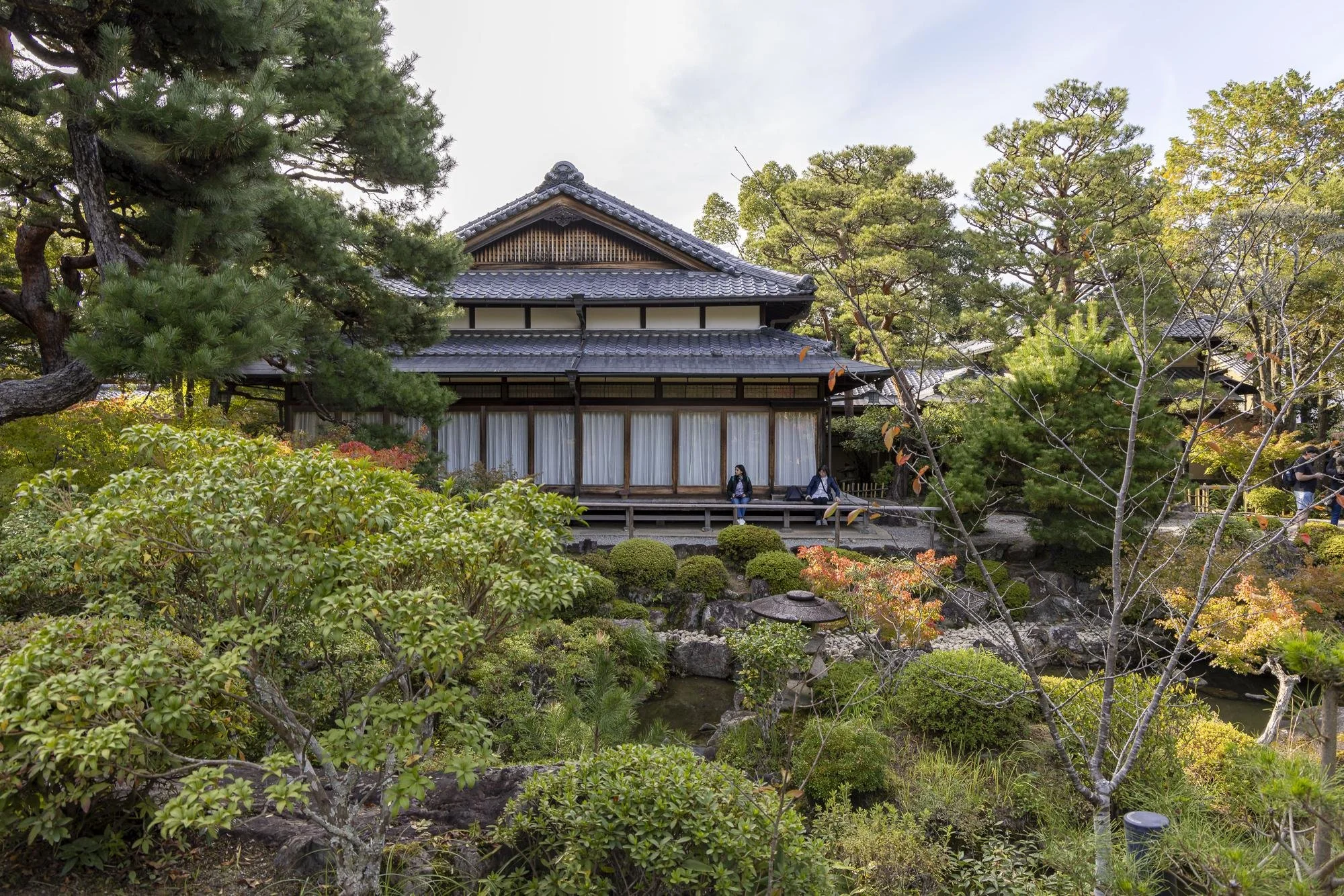 Traditional Japanese wooden house surrounded by lush greenery and trees, with a small pond in the garden.