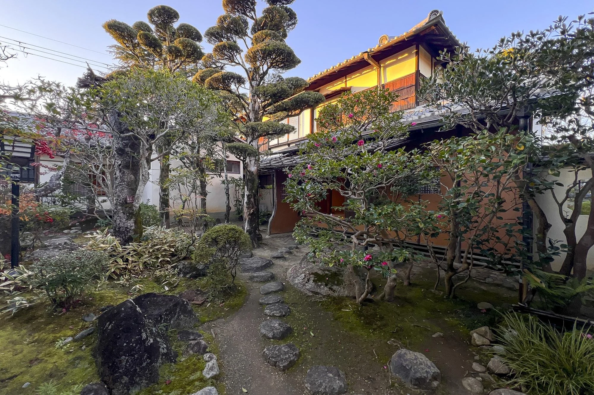 Traditional Japanese garden with a stone pathway, trees, shrubs, moss, and a wooden house with shoji door in the background.