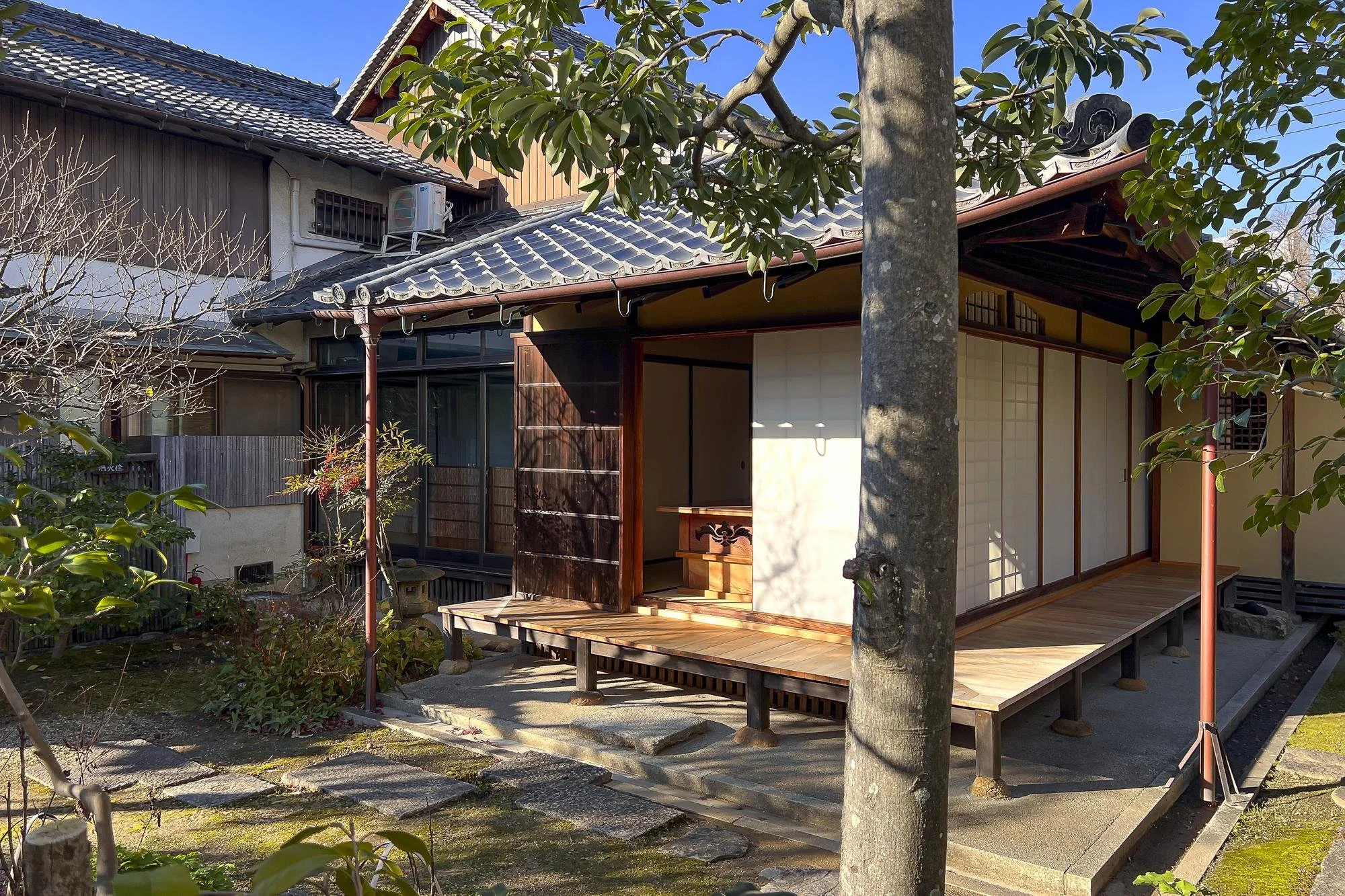 A traditional Japanese house with a wooden porch, shoji sliding doors, a tiled roof, and a garden with trees and stones.