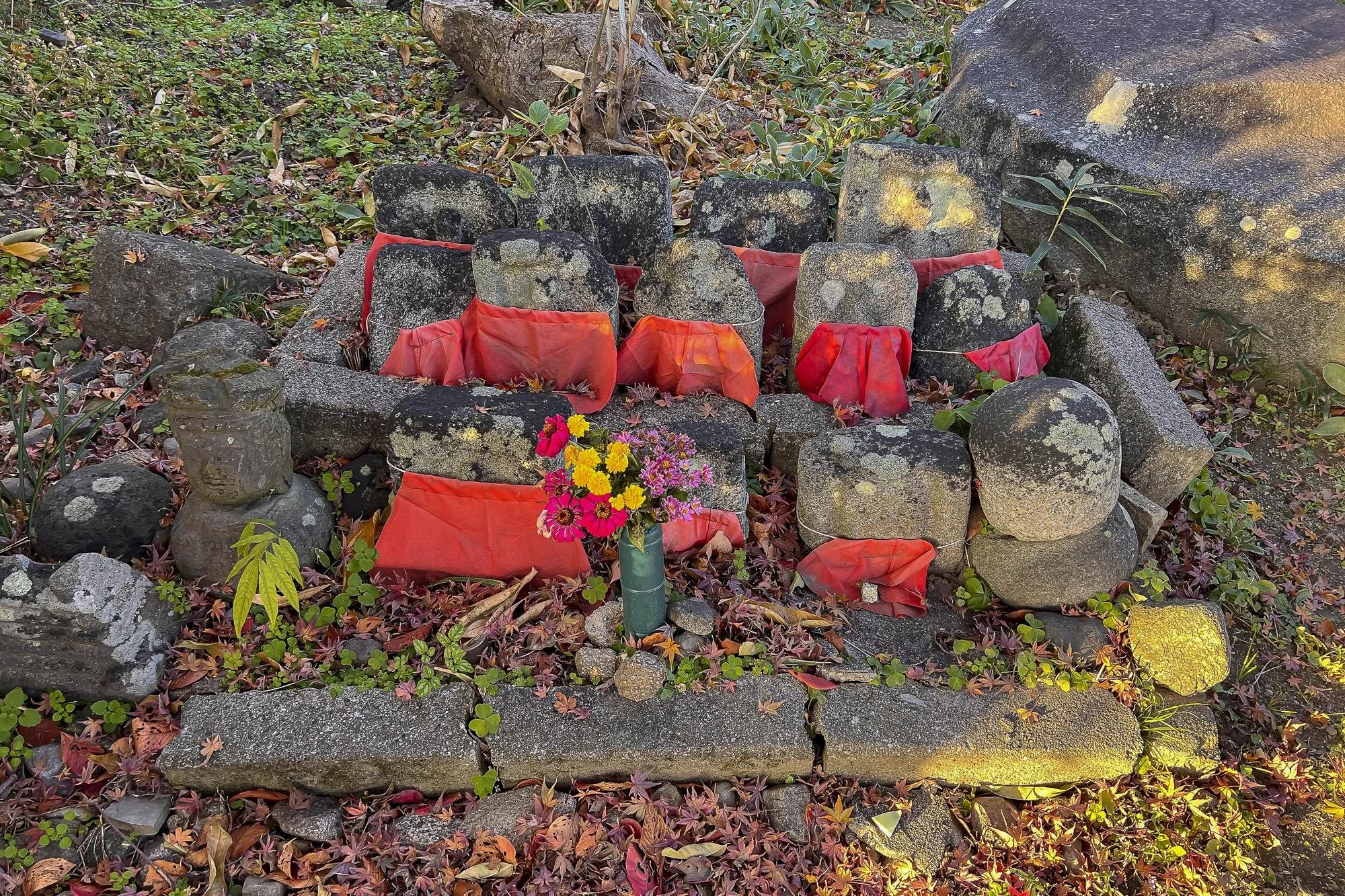 A small stone shrine with weathered rocks, decorated with red fabric coverings and a flower arrangement at the center, surrounded by fallen leaves and greenery.