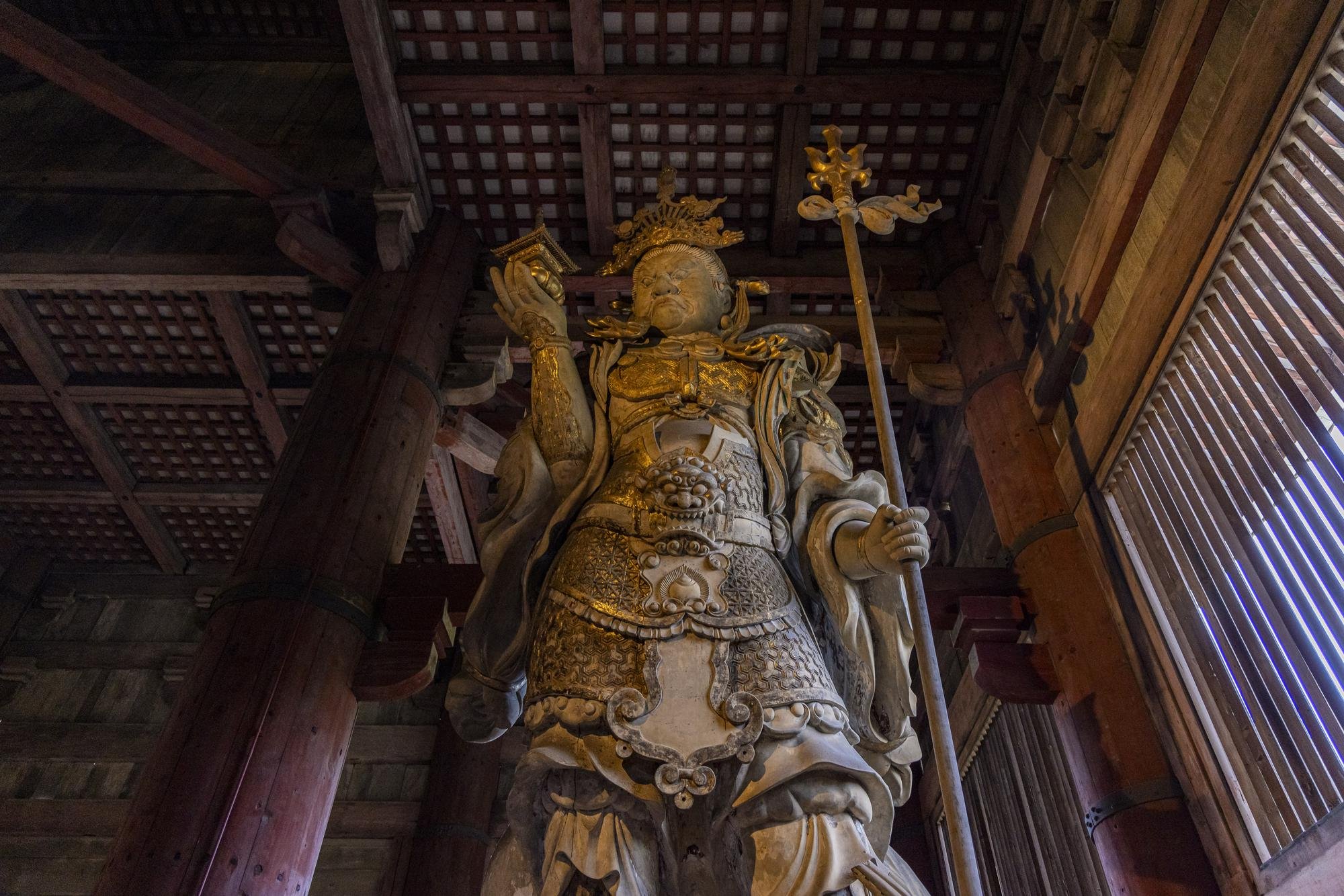 A tall, ornate wooden statue of a warrior or deity inside a temple with traditional wooden architecture, visible wooden beams and lattice windows.