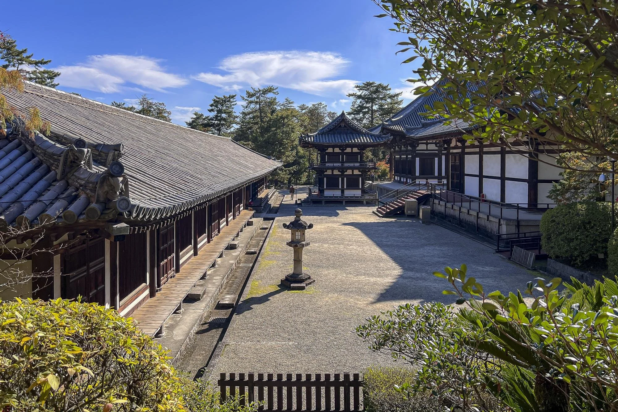 Traditional Japanese temple structures with tiled roofs, stone lantern, and surrounding greenery under a blue sky.