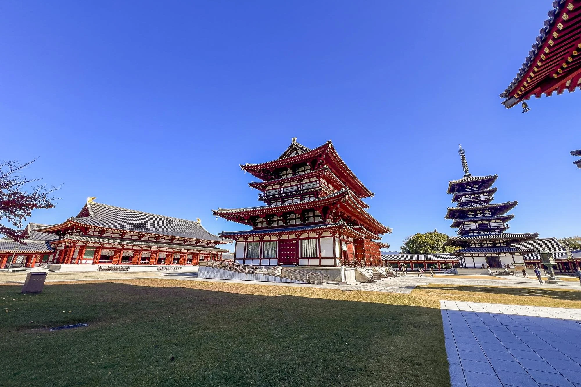 Traditional Japanese temple buildings with red and white color schemes, pagoda-style towers, a clear blue sky, and a grassy courtyard in the foreground.