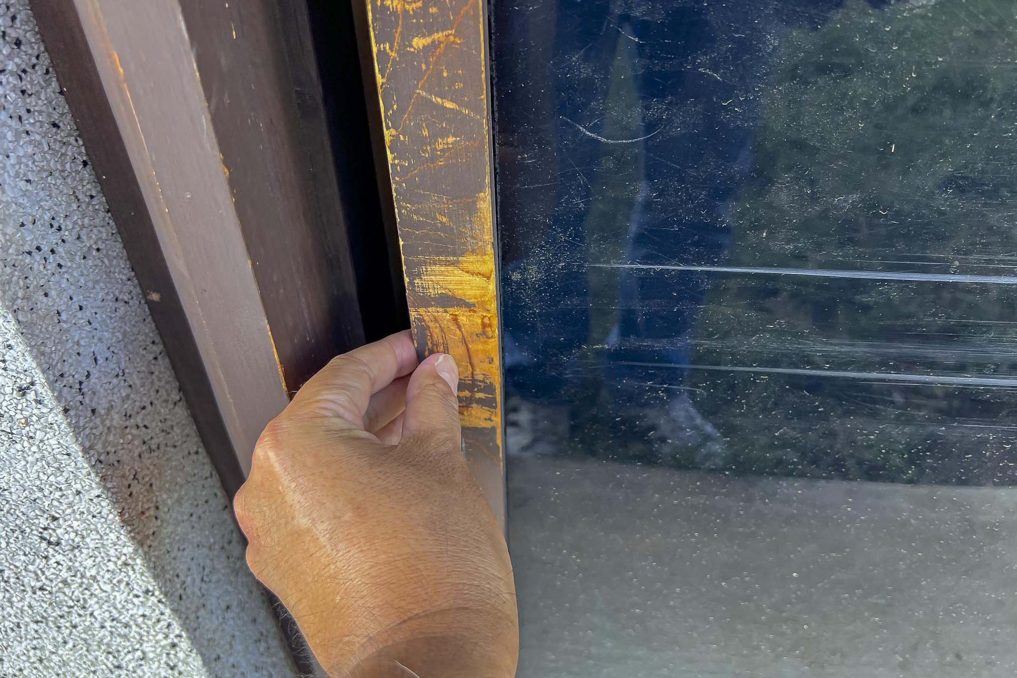 Close-up of a hand lifting the corner of a worn wooden frame to reveal a dark glass panel, with a textured light-colored wall in the background.