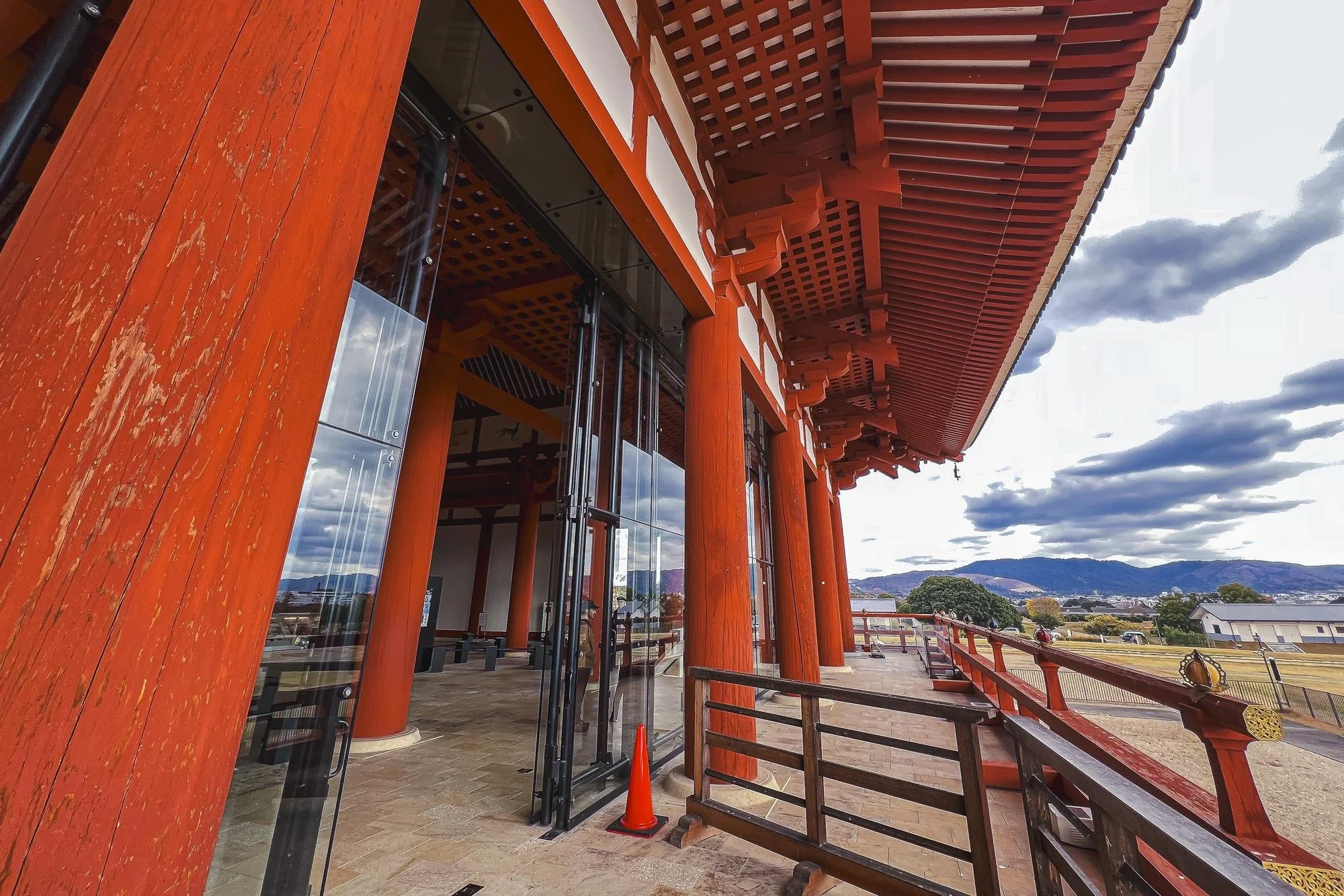 Traditional Japanese building with red wooden beams, pillars, and detailed roof structure, set against a mountainous landscape with partly cloudy sky.