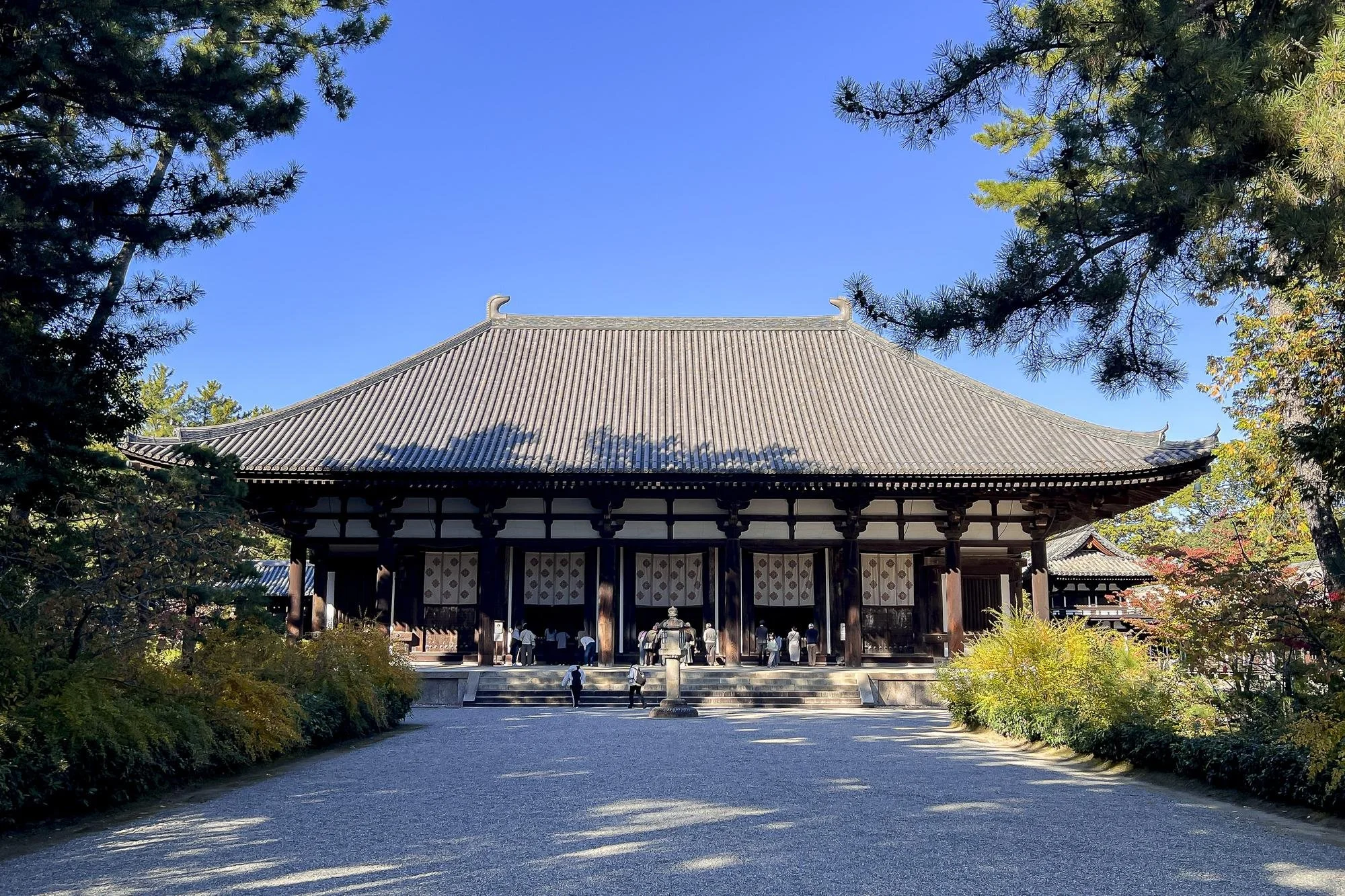 Traditional Japanese temple with wooden structure and tiled roof, surrounded by trees and shrubs under a clear blue sky.