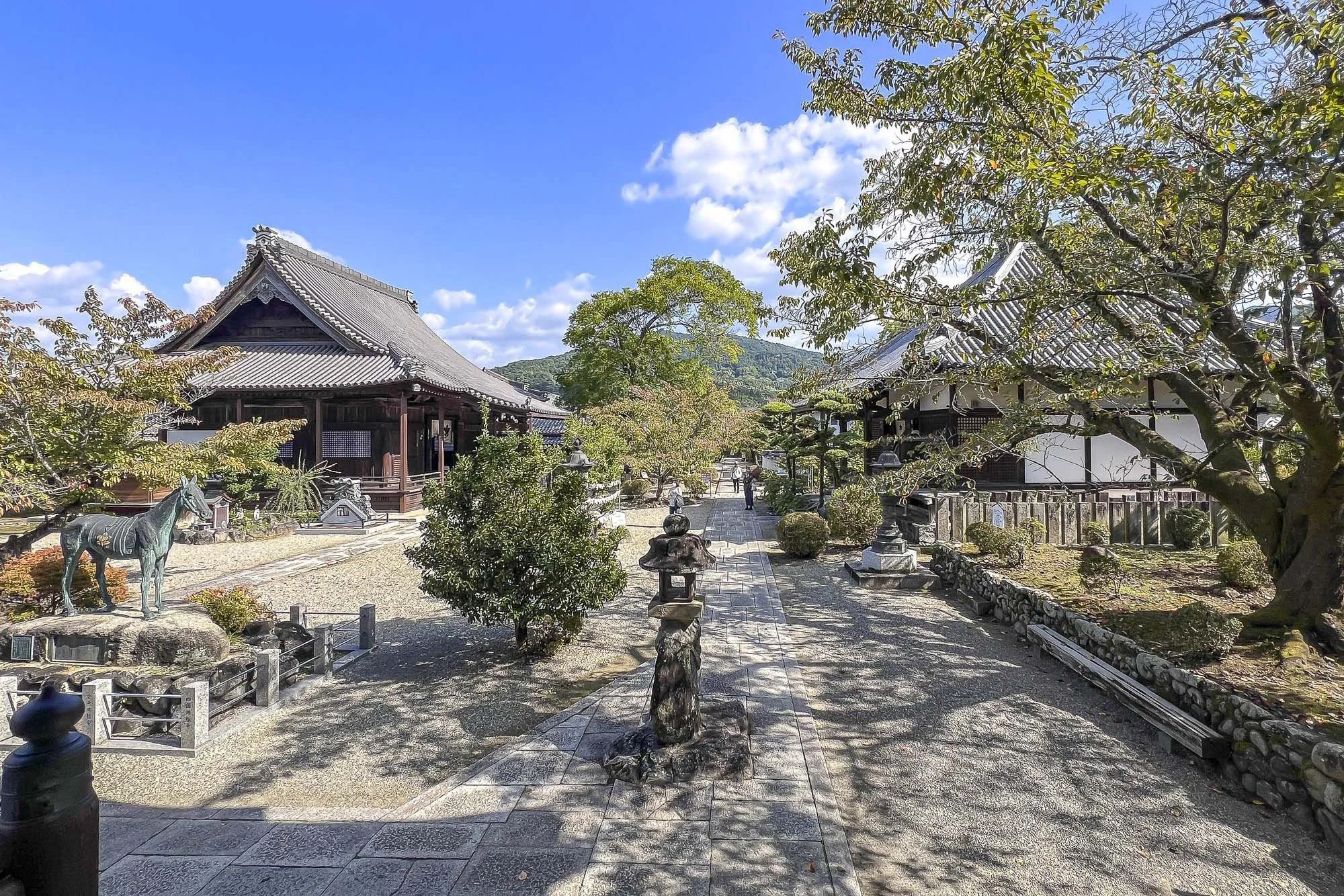 Traditional Japanese temple with wooden buildings, stone lanterns, and a horse statue, surrounded by trees and a clear blue sky.