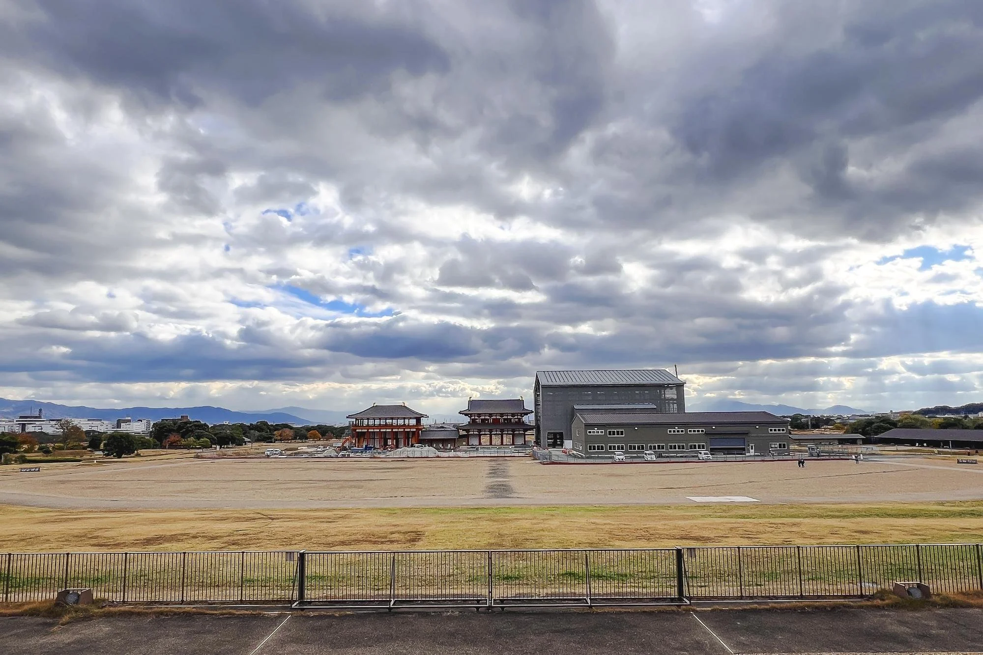 Traditional Japanese building structures in an open, sandy area under cloudy sky, with mountains in the distant background and a black fence in the foreground.