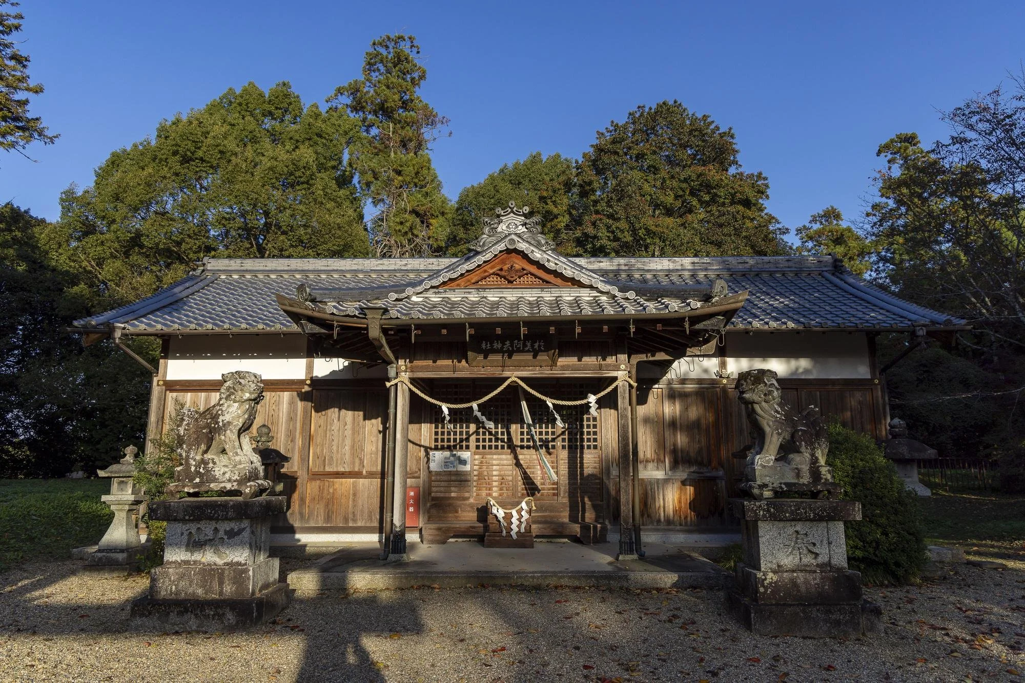 A traditional Japanese Shinto shrine with a wooden structure, tiled roof, and two stone guardian statues outside, surrounded by trees.