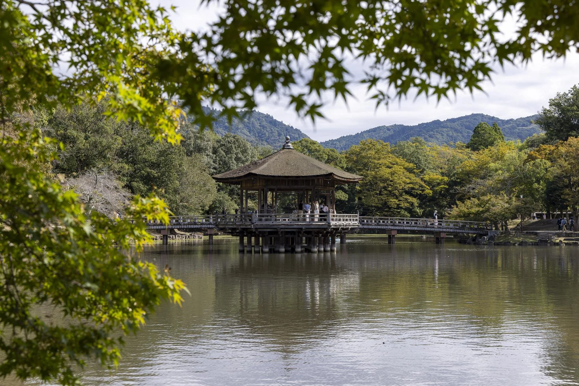 A traditional Japanese gazebo on a lake, surrounded by lush green trees and distant mountains, with people walking along a wooden bridge.