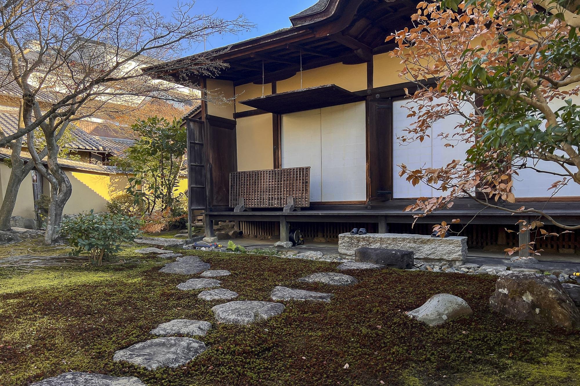 Traditional Japanese house with sliding shoji doors, wooden porch, and surrounding Japanese garden with moss and rocks.