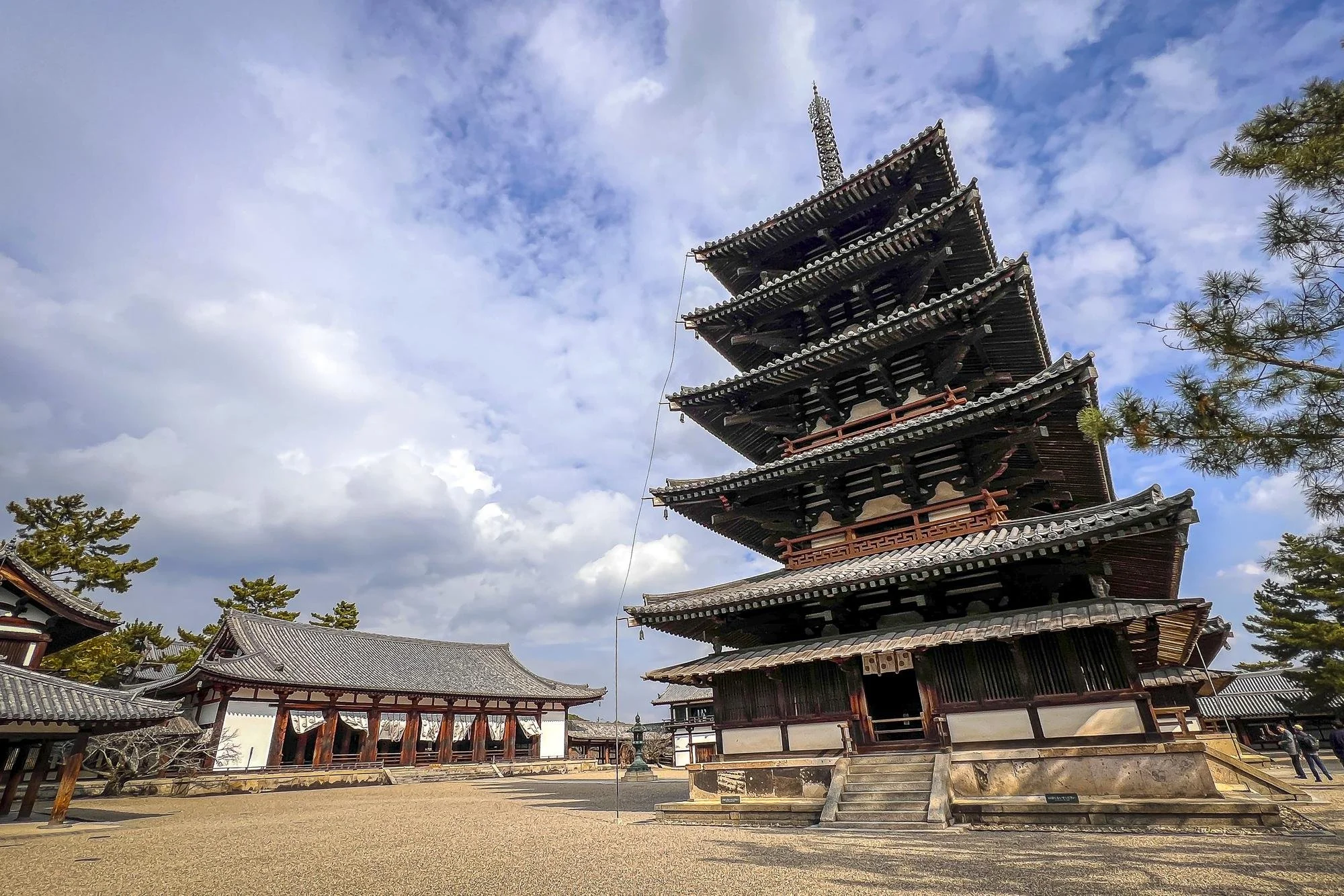 A traditional multi-story pagoda with curved eaves and wooden construction, set in an open courtyard with trees and other wooden buildings around.