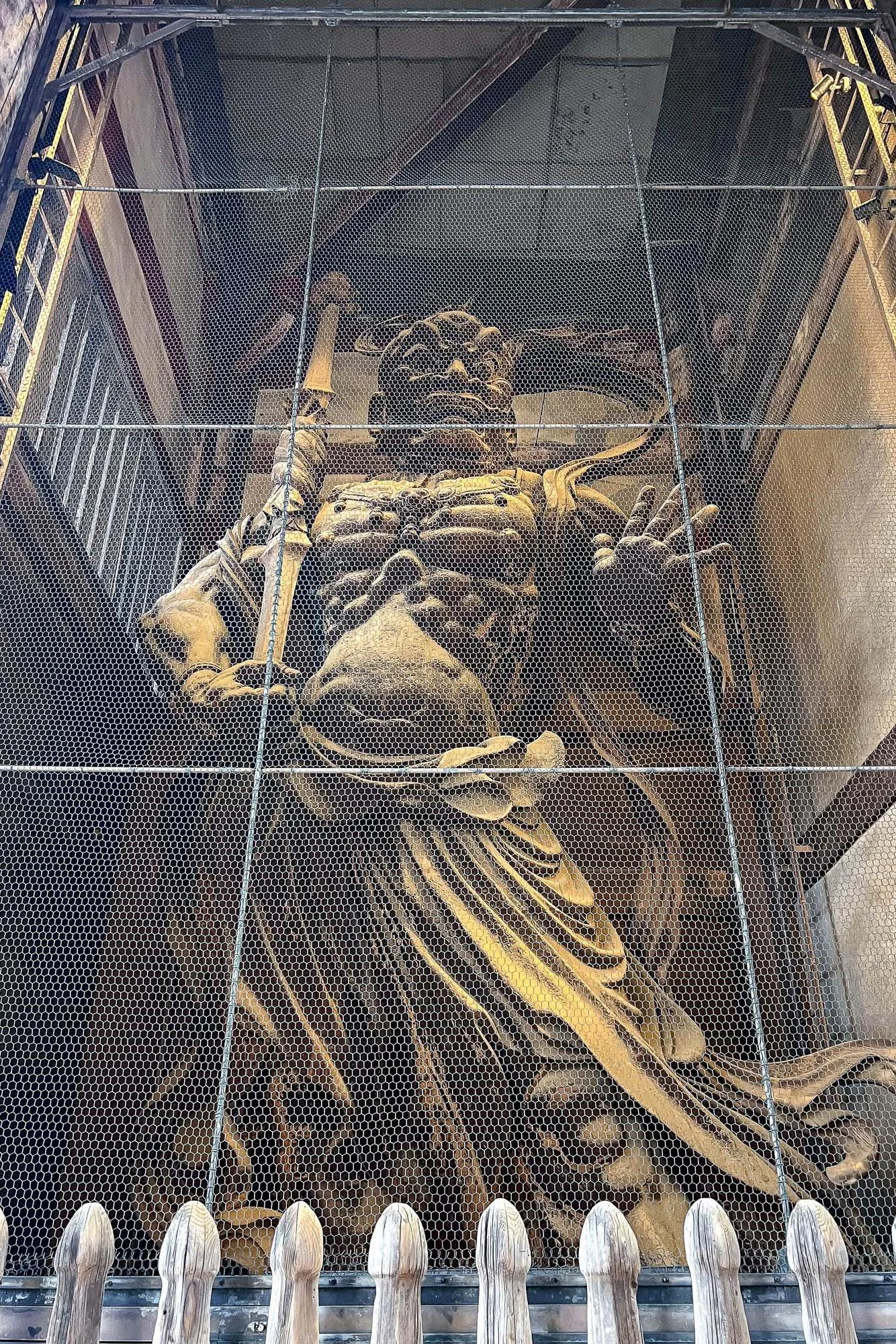 Large statue of a warrior with a headdress, holding a spear, surrounded by a metal mesh cage.