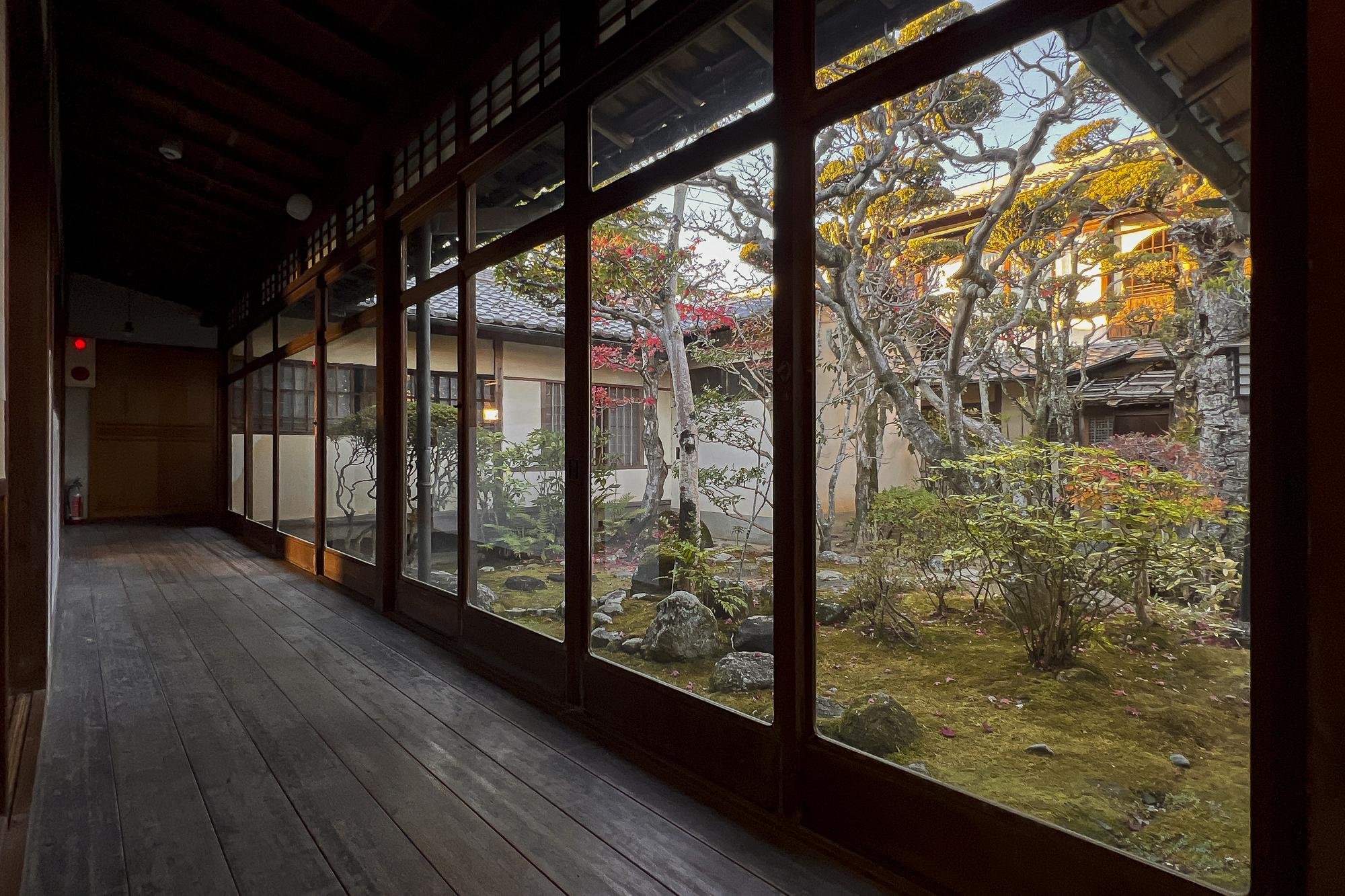 View of a serene Japanese garden through sliding glass doors with wooden frames, showing trees and bushes with autumn foliage and a traditional building in the background.