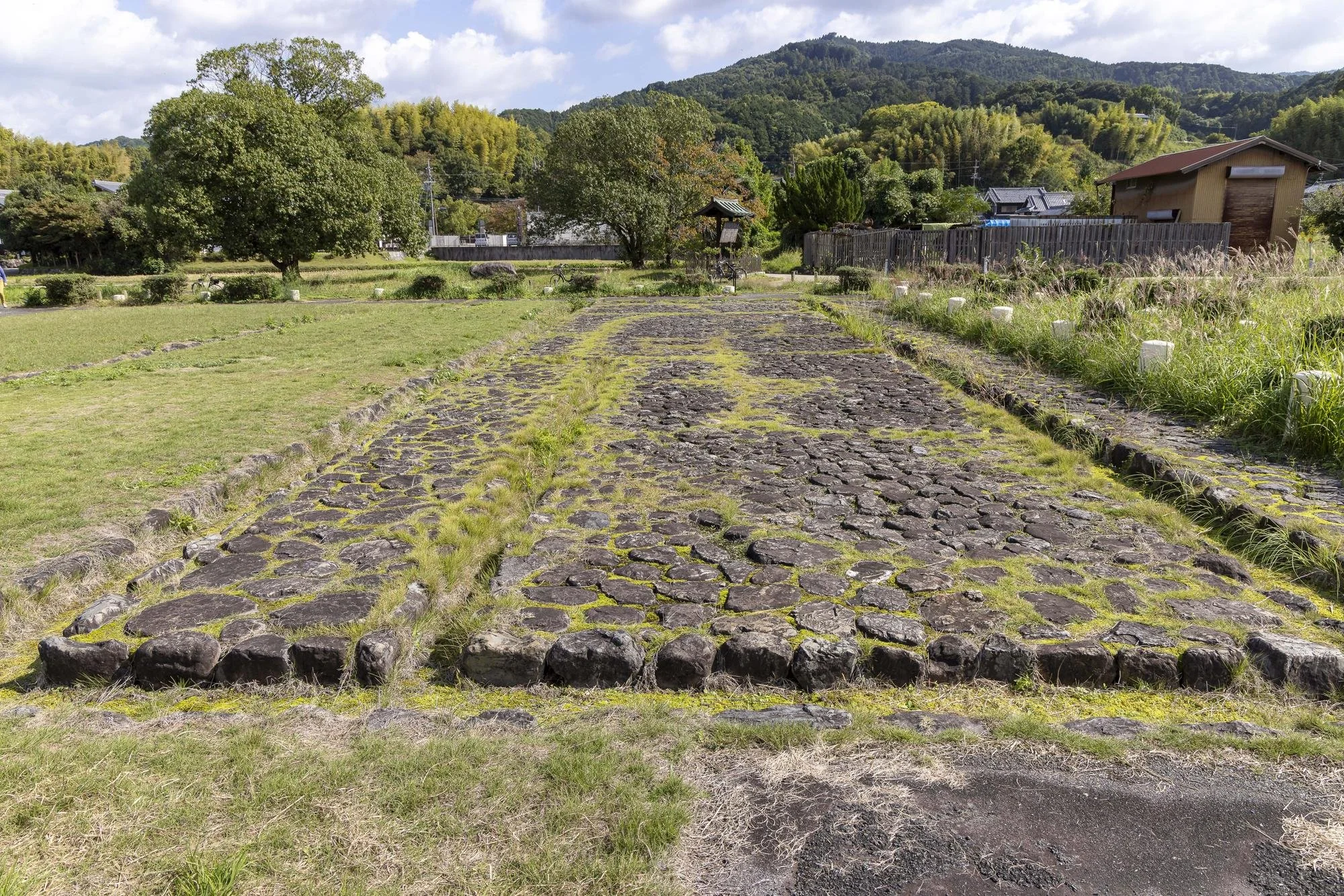 Asuka Palace Site: Ancient stone pathway in a grassy outdoor area with trees, buildings, and mountains in the background on a partly cloudy day.