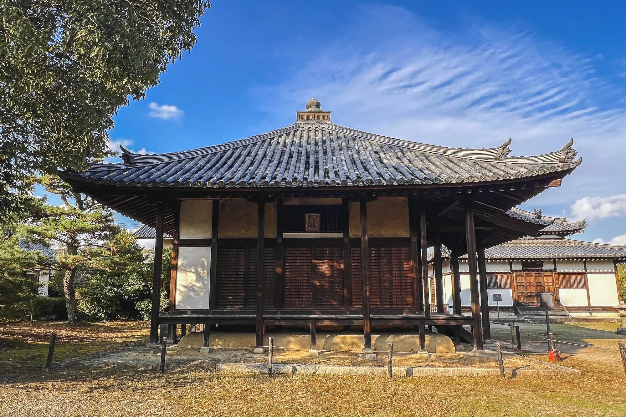 A traditional wooden Japanese temple with a tiled, curved roof surrounded by trees and an open yard, under a partly cloudy sky.