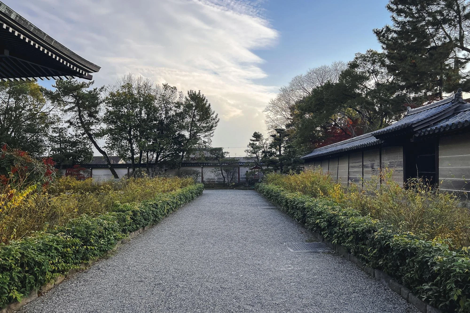 A peaceful gravel pathway lined with green bushes on each side, leading through a traditional Japanese setting with trees and buildings with tiled roofs under a partly cloudy sky.