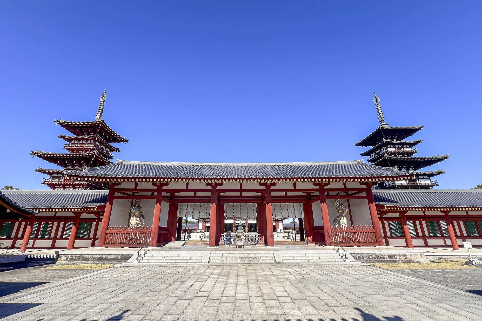 A traditional Japanese temple with red and white walls, two multi-tiered pagoda towers on either side, and a spacious paved courtyard under a clear blue sky.
