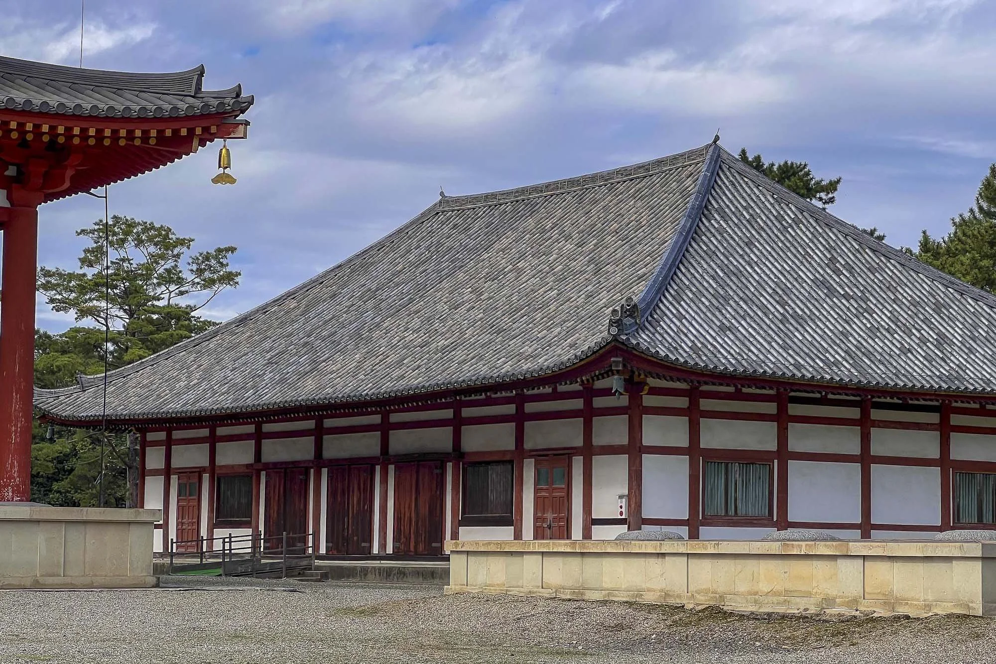 Traditional Japanese temple with a large, curved tiled roof, white walls with red wooden framing, and small windows, set against a backdrop of trees and a cloudy sky.