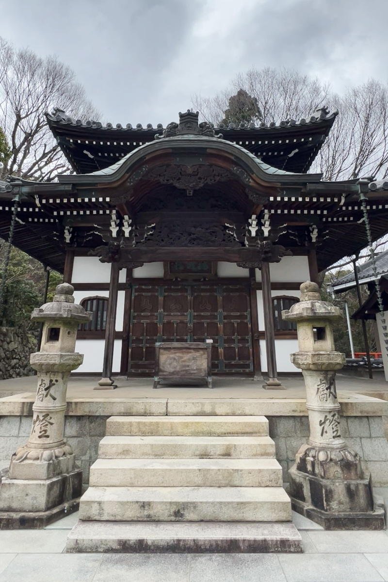 A traditional Japanese temple with a wooden structure, dark roof, stone lanterns, and stone steps leading up to the entrance, surrounded by leafless trees under a cloudy sky.