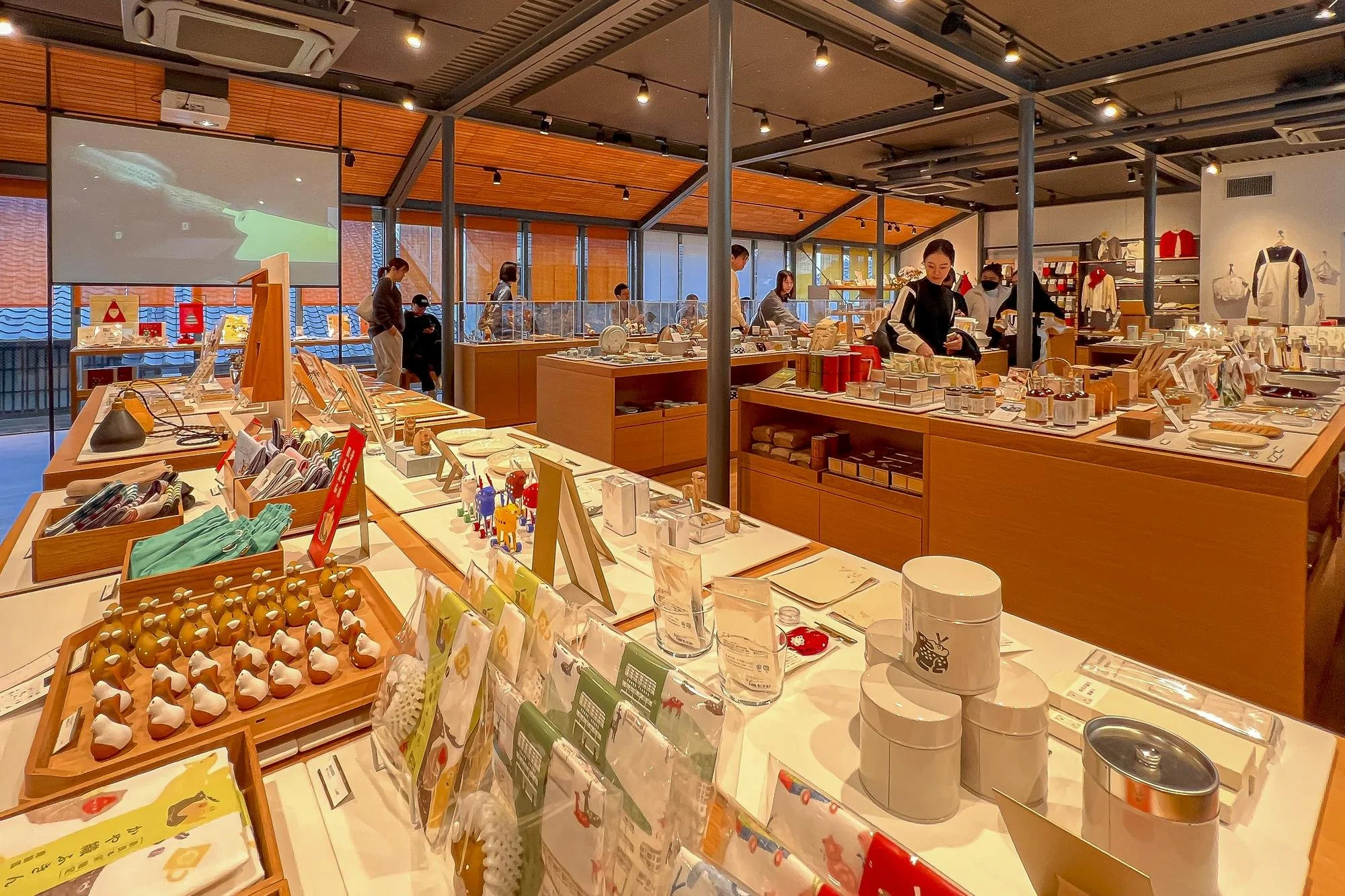 Inside of a retail store displaying various small items, such as toys, accessories, and household goods, arranged on wooden tables. There are multiple customers and staff members shopping and working, with a large window and ceiling lights illuminati