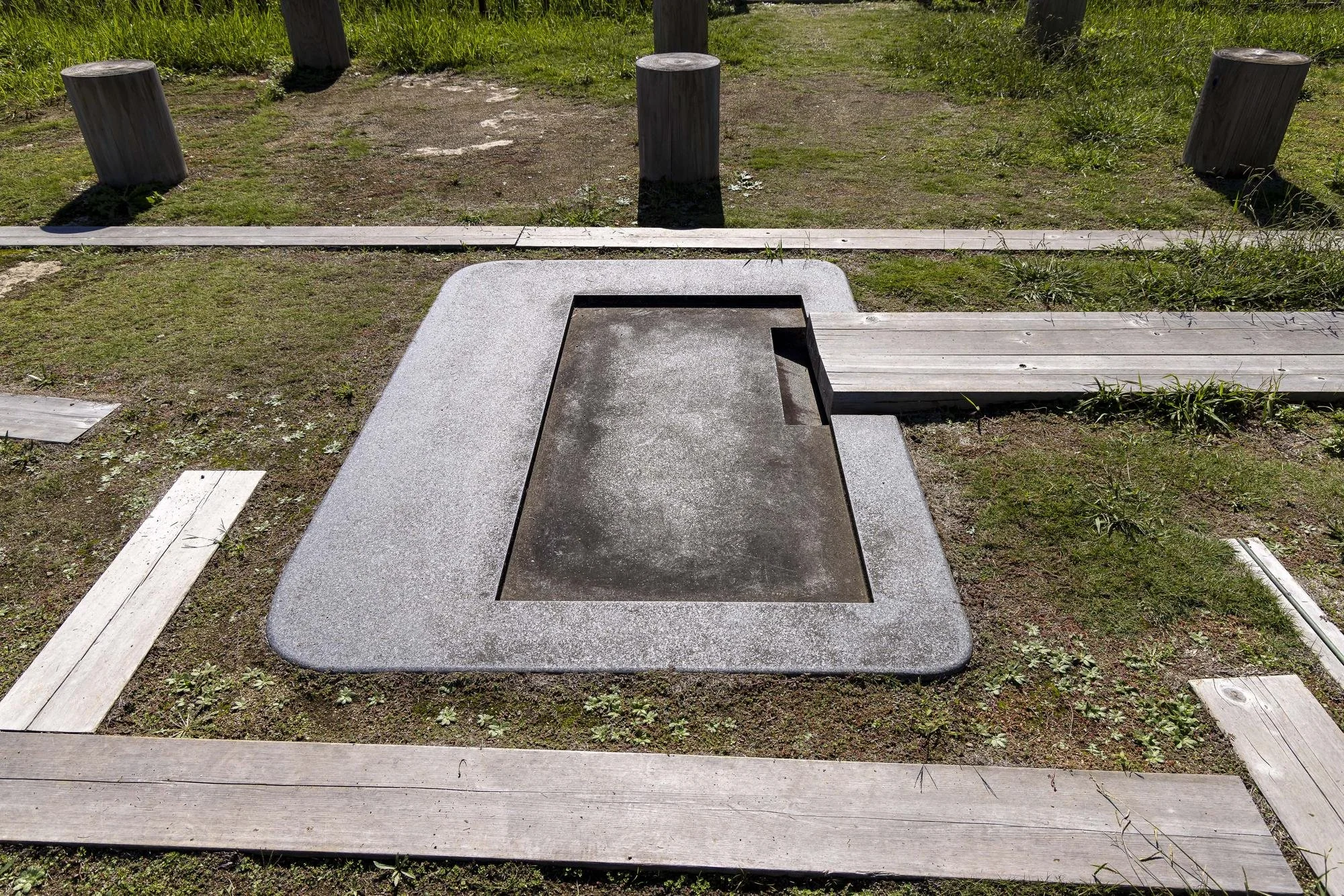 A disc golf basket with a metal basket and wooden obstructing bars, situated on a patch of dirt and grass with a wooden walking trail and stumps surrounding it.