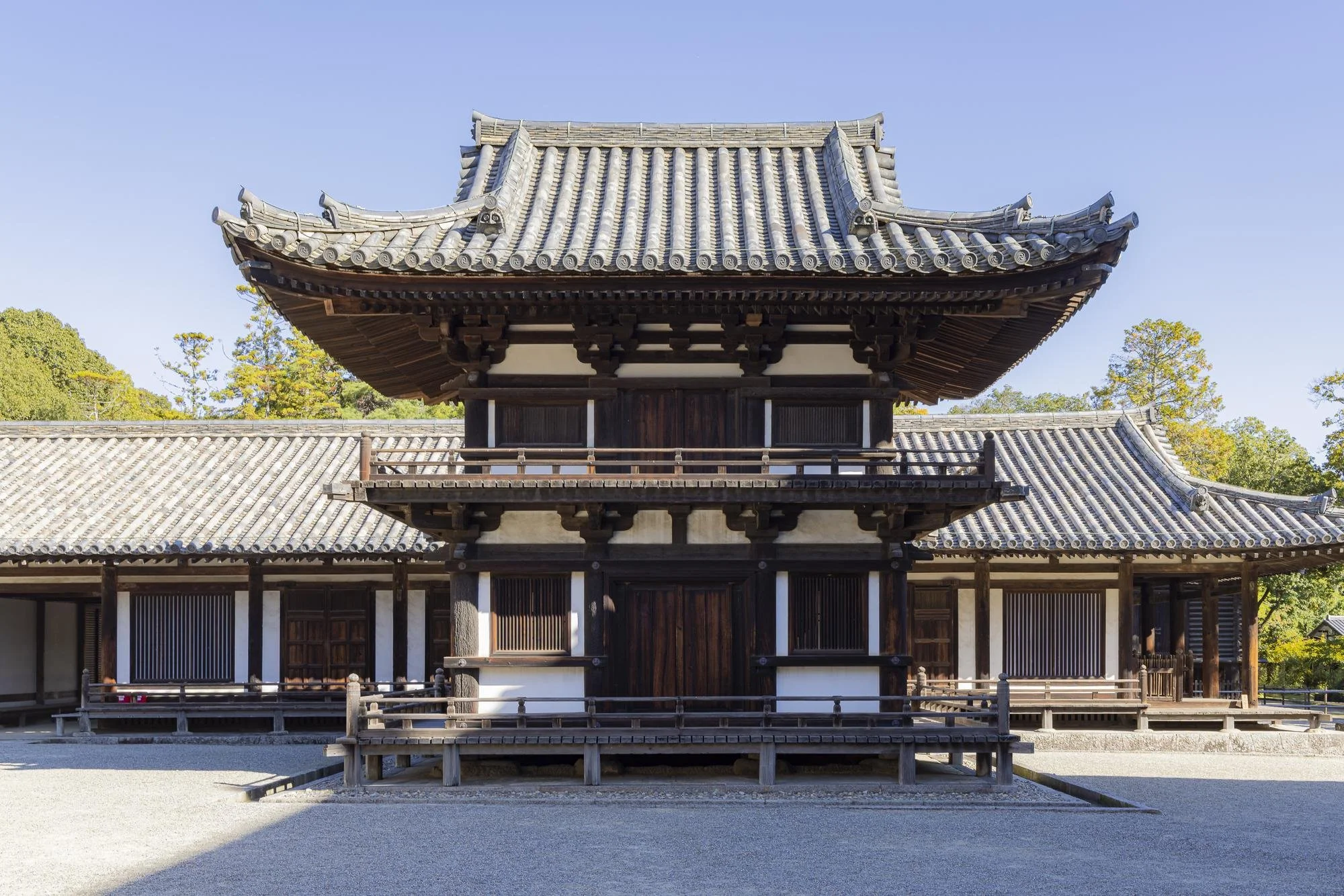 A traditional Japanese temple with a curved tiled roof and wooden structure, surrounded by trees and a gravel courtyard.