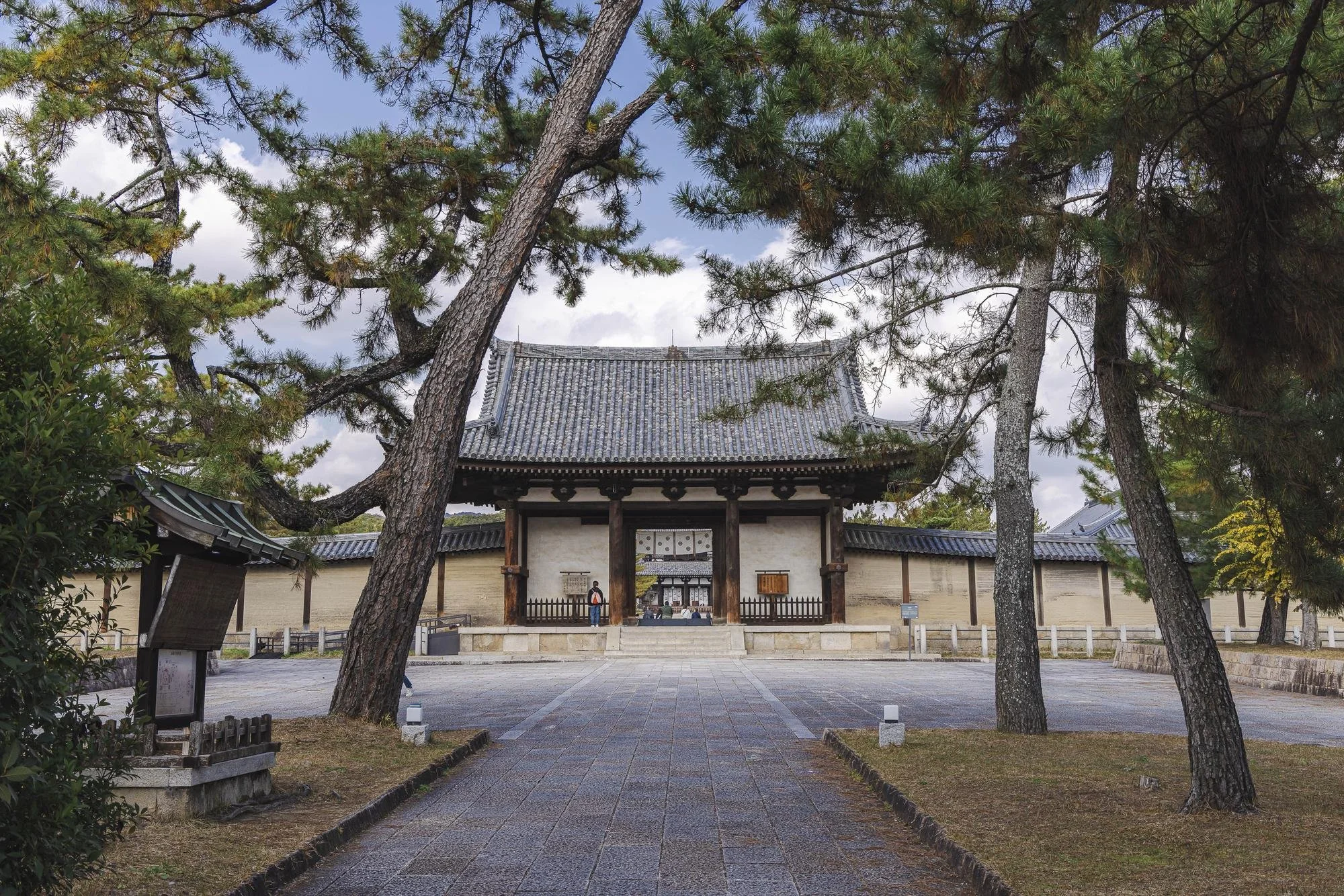 A traditional Japanese temple entrance, framed by tall trees and a stone pathway leading to the gate.