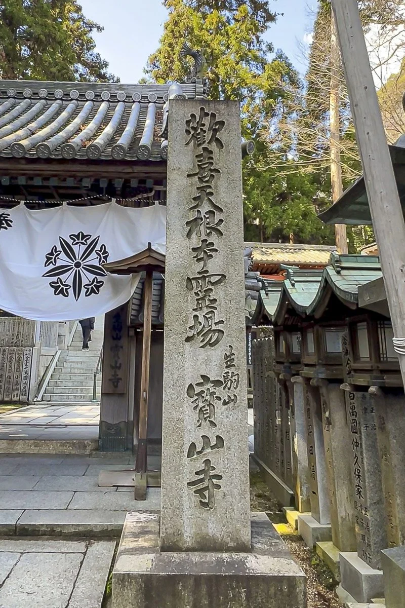 A stone monument with Japanese inscriptions at a traditional Japanese temple, with trees and a building with a tiled roof in the background.