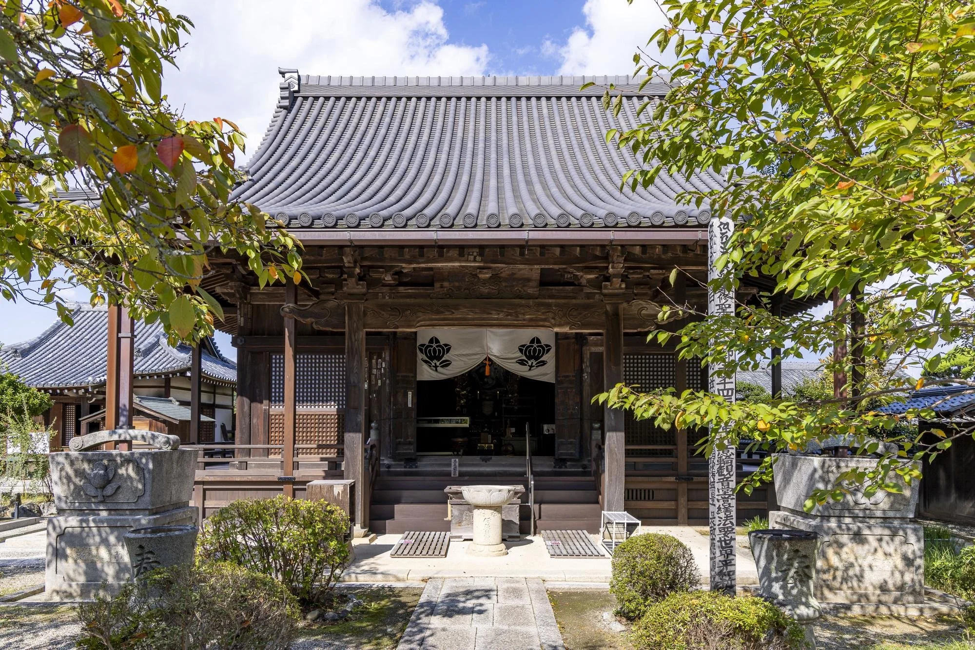 A traditional Japanese temple with a wooden structure, tiled roof, and stone ornaments, surrounded by greenery under a partly cloudy sky.
