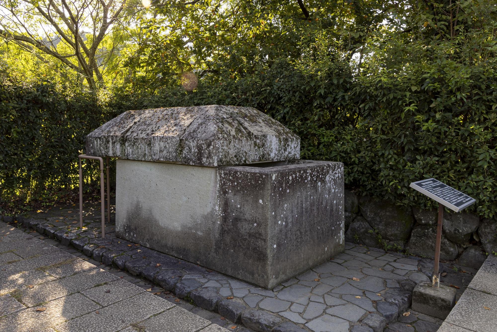 Large stone monument with a sloped top, surrounded by a paved walkway and a metal railing, with bushes and trees in the background, and an informational plaque on the right side.