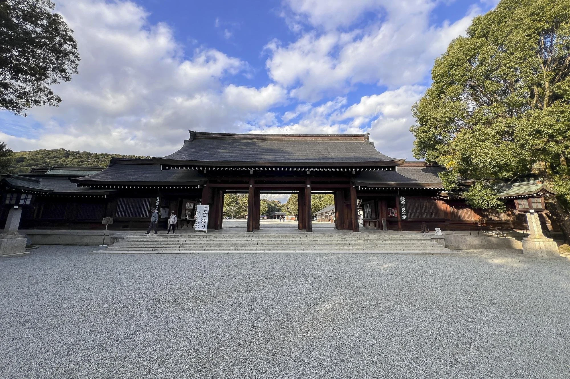 Traditional Japanese temple entrance with a large wooden gate, steps, and surrounding trees under a partly cloudy sky.