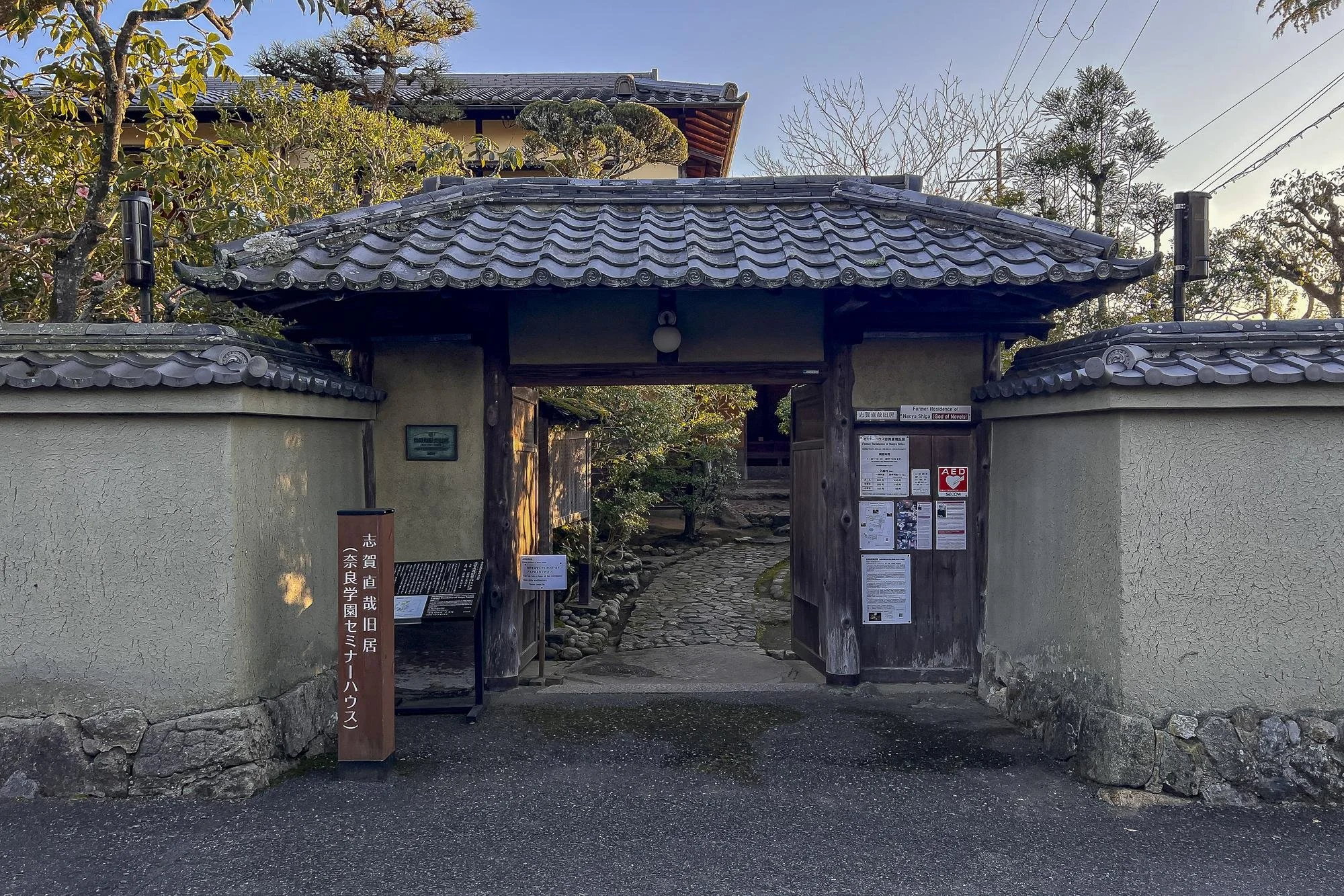 The image shows a traditional Japanese entrance gate with a tiled roof, wooden frame, and stone walls. There is a pathway leading inside with trees and foliage in the background. Various signs and notices are posted near the gate.